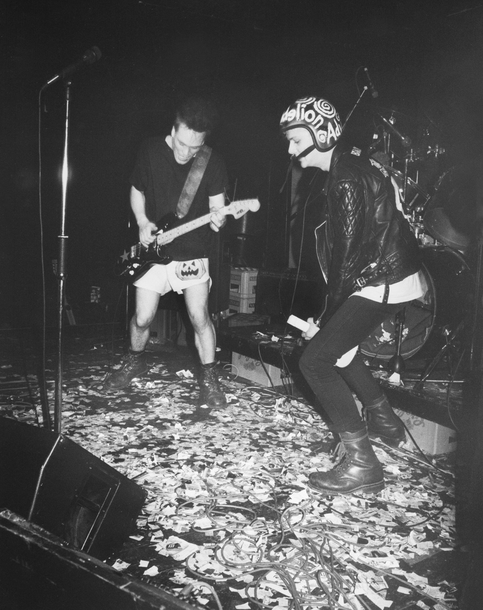 a band on stage, one member on the left is wearing white shorts with a pumpkin on them and a black t-shirt. they play a guitar enthusiastically. a singer wearing skinny black jeans, a leather jacket and a motorbike helmet. the stage is covered in torn paper.