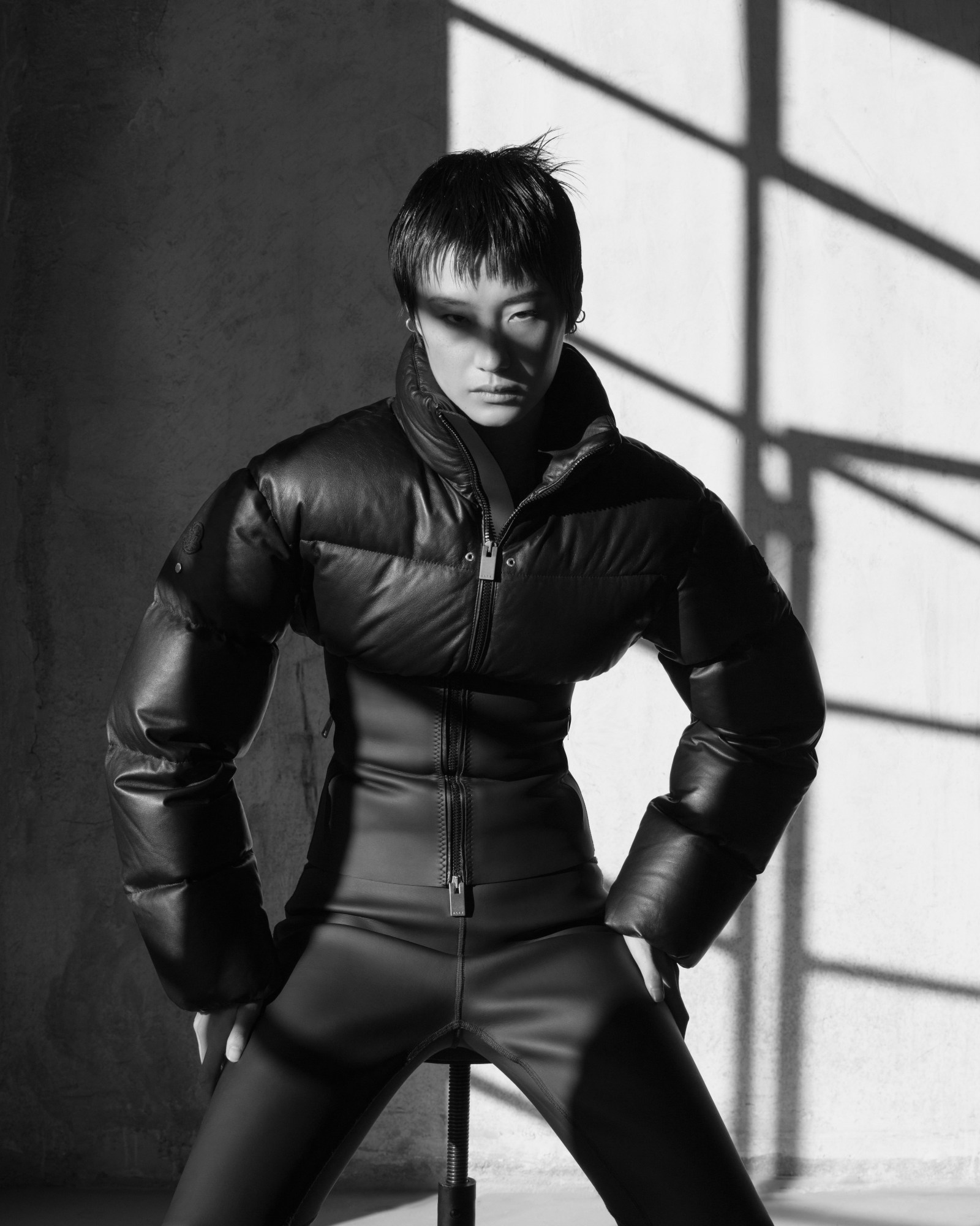 a model wearing a puffer jacket, cinched at the waist, poses in a shadowy outdoor space