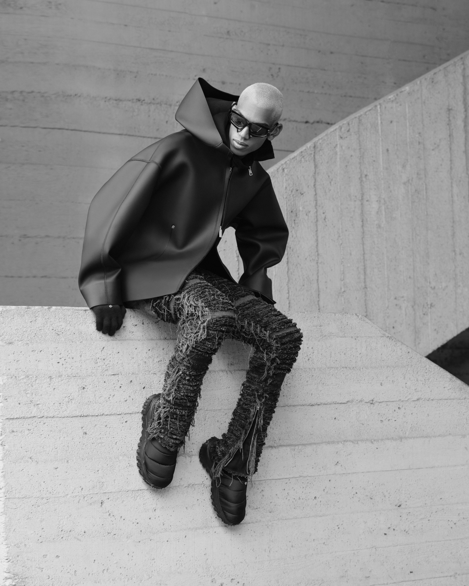 a model wearing messy knit trousers and a neoprene type waterproof jacket poses on a concrete wall