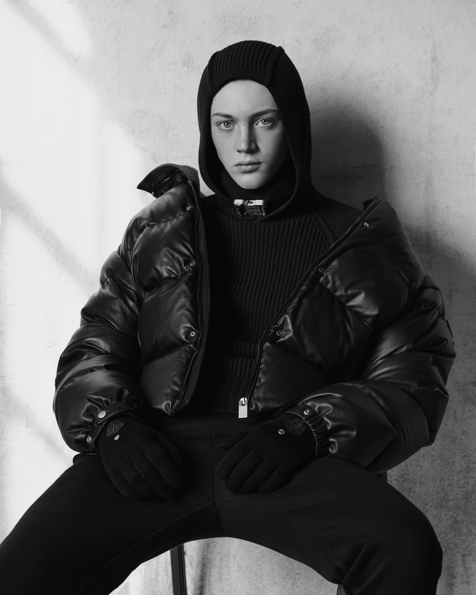 a model wearing a hooded knit slouches on a chair in a black puffer jacket