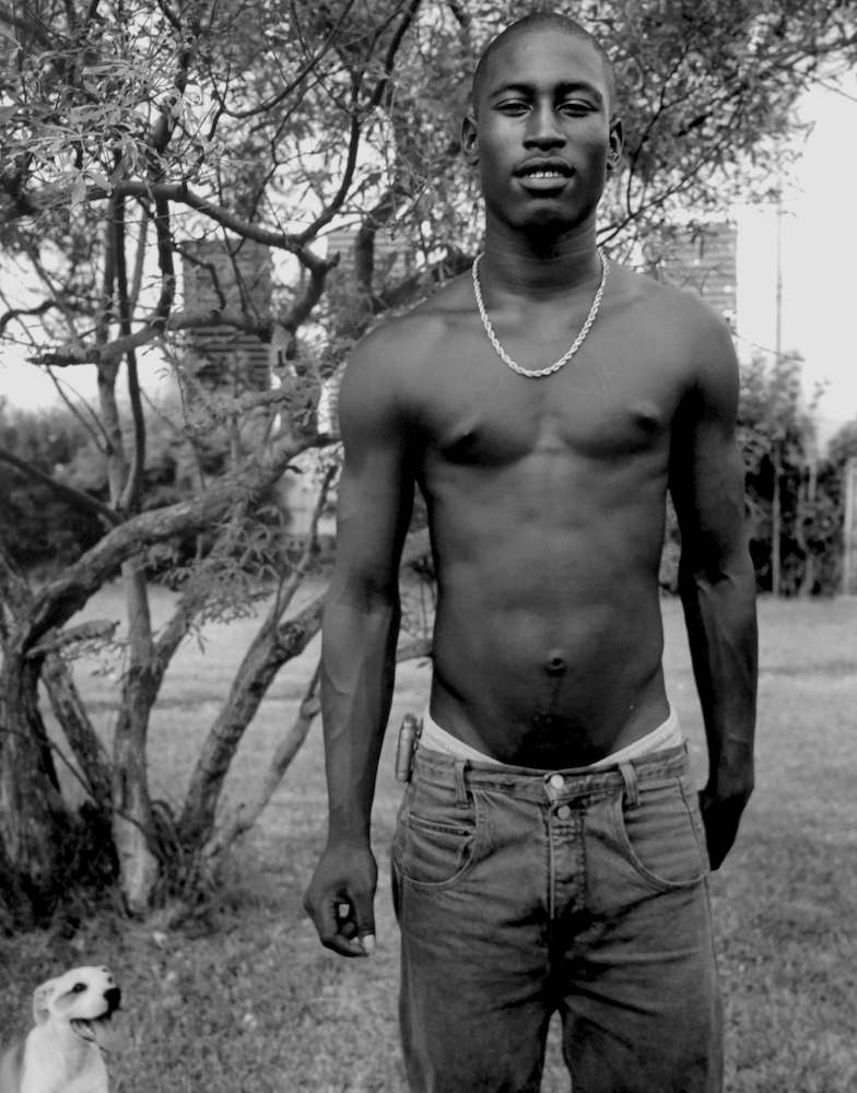 A white dog and a shirtless young man wearing a gold necklace in front of a tree