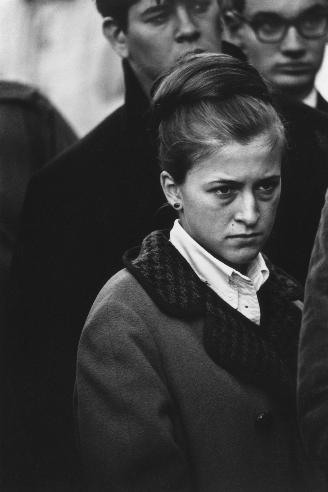 a woman wearing a black coat stares into the distance with a serious expression