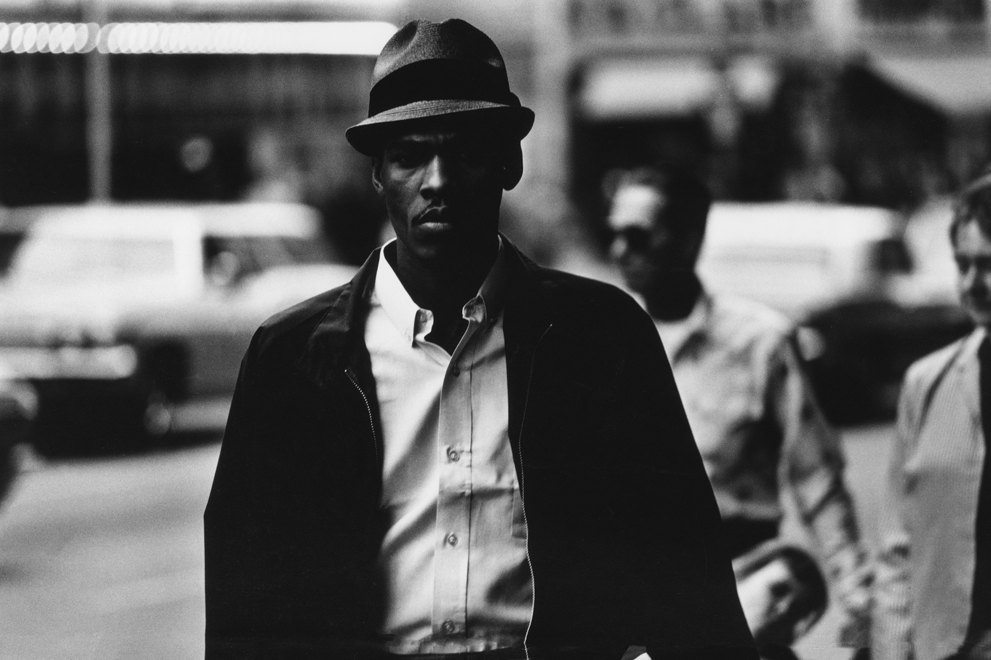 a man wearing a hat walking down the street stares into the camera