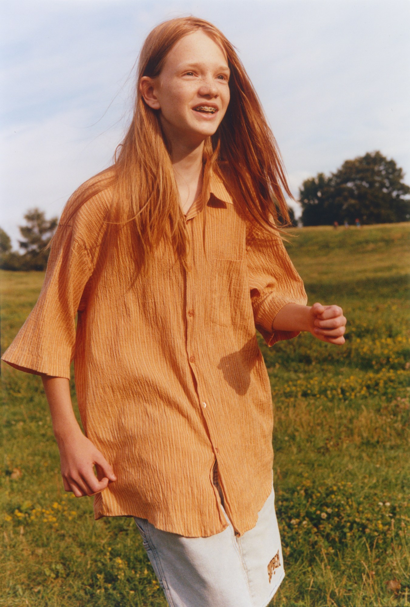 A teenager in a baggy orange shirt and long orange hair smiling in a field