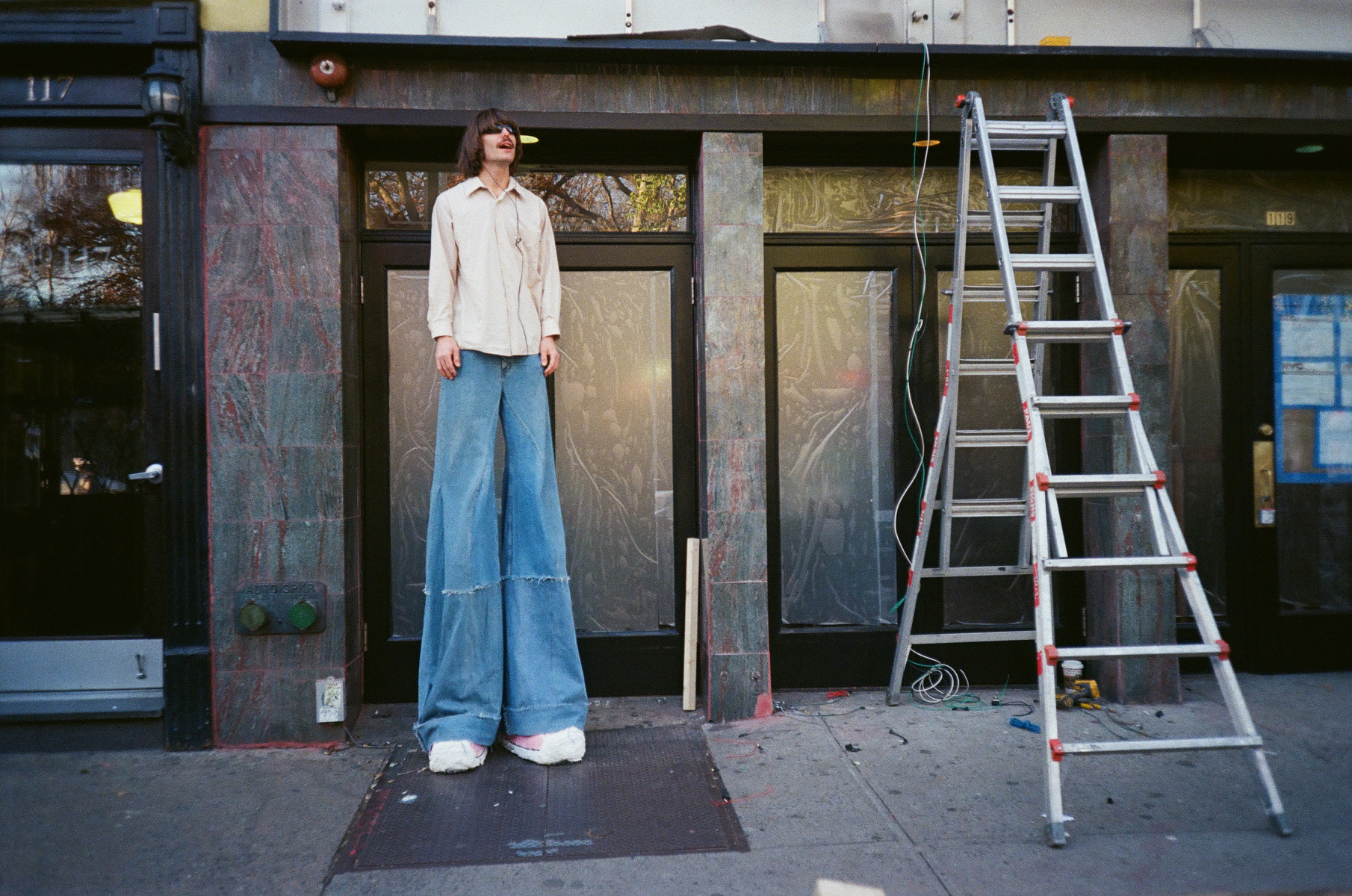 a man with disproportionately large legs in blue bellbottom jeans and a beige shirt wearing stilts standing next to a ladder