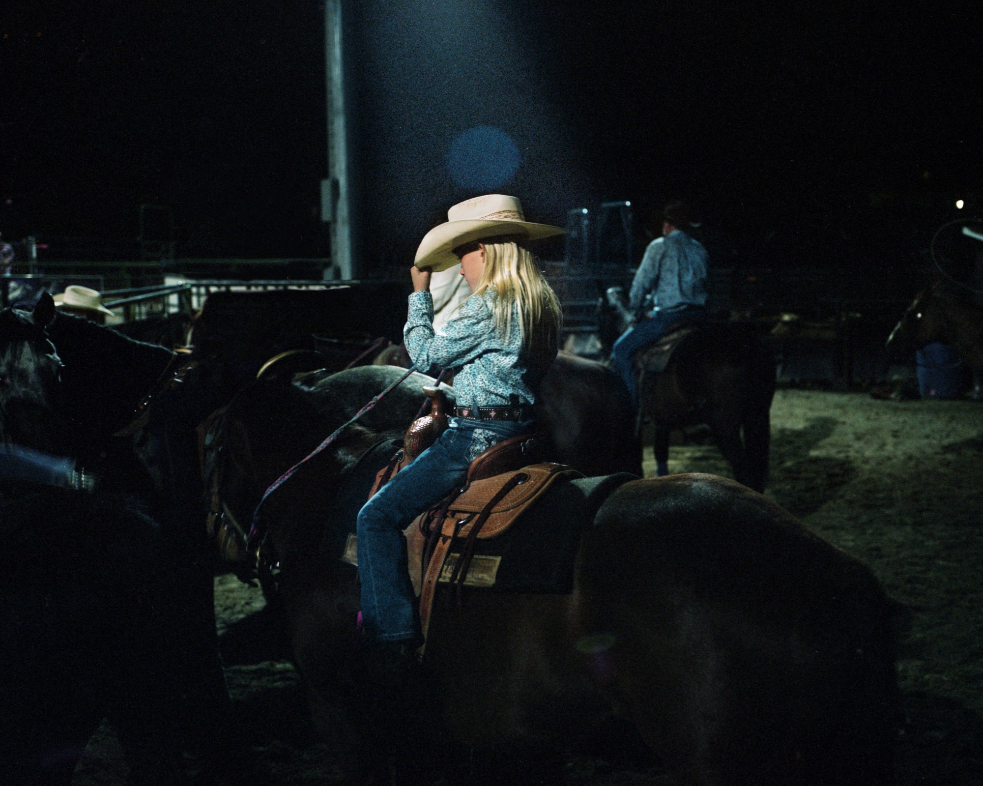 a blonde girl sitting on top of a horse at night wearing jeans, floral shirt and cowboy hat