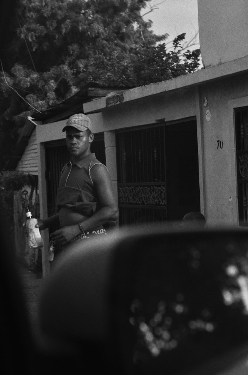 black-and-white image of a man holding alcohol in a brown paper bag with car mirror in foreground