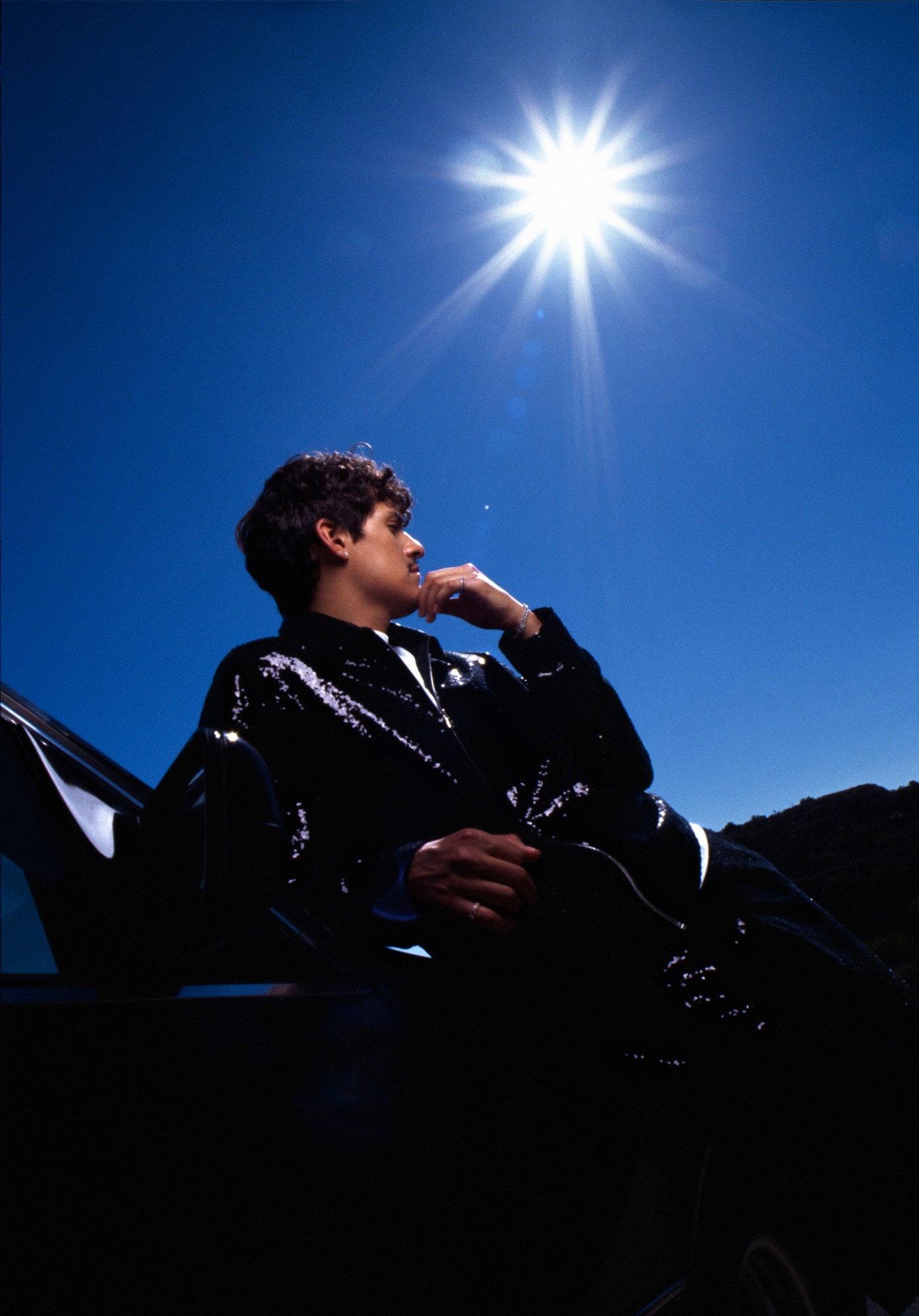 musician omar apollo dressed in black vinyl and leaning across a car, looking off into the distance in front of a blue sky