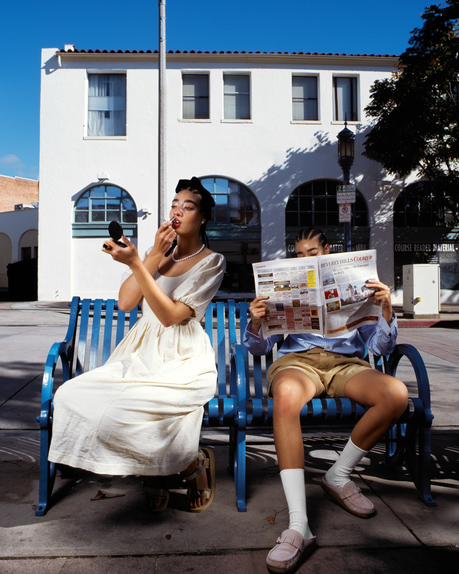 a couple sit beside each other on a blue bench, one touching up her make up and the other reading the newspaper