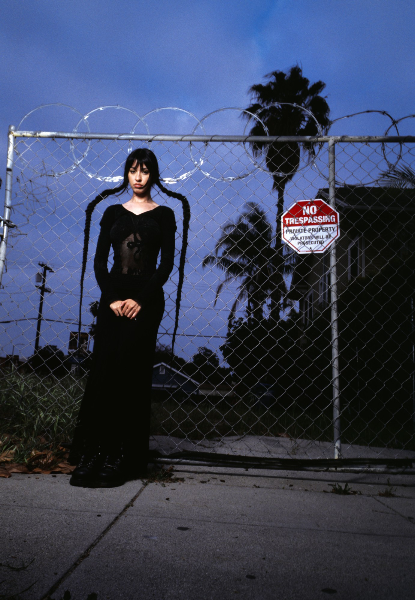 a model in a long black dress with two long plaits stands in front of a chain link fence and a 'no trespassing' sign at dusk