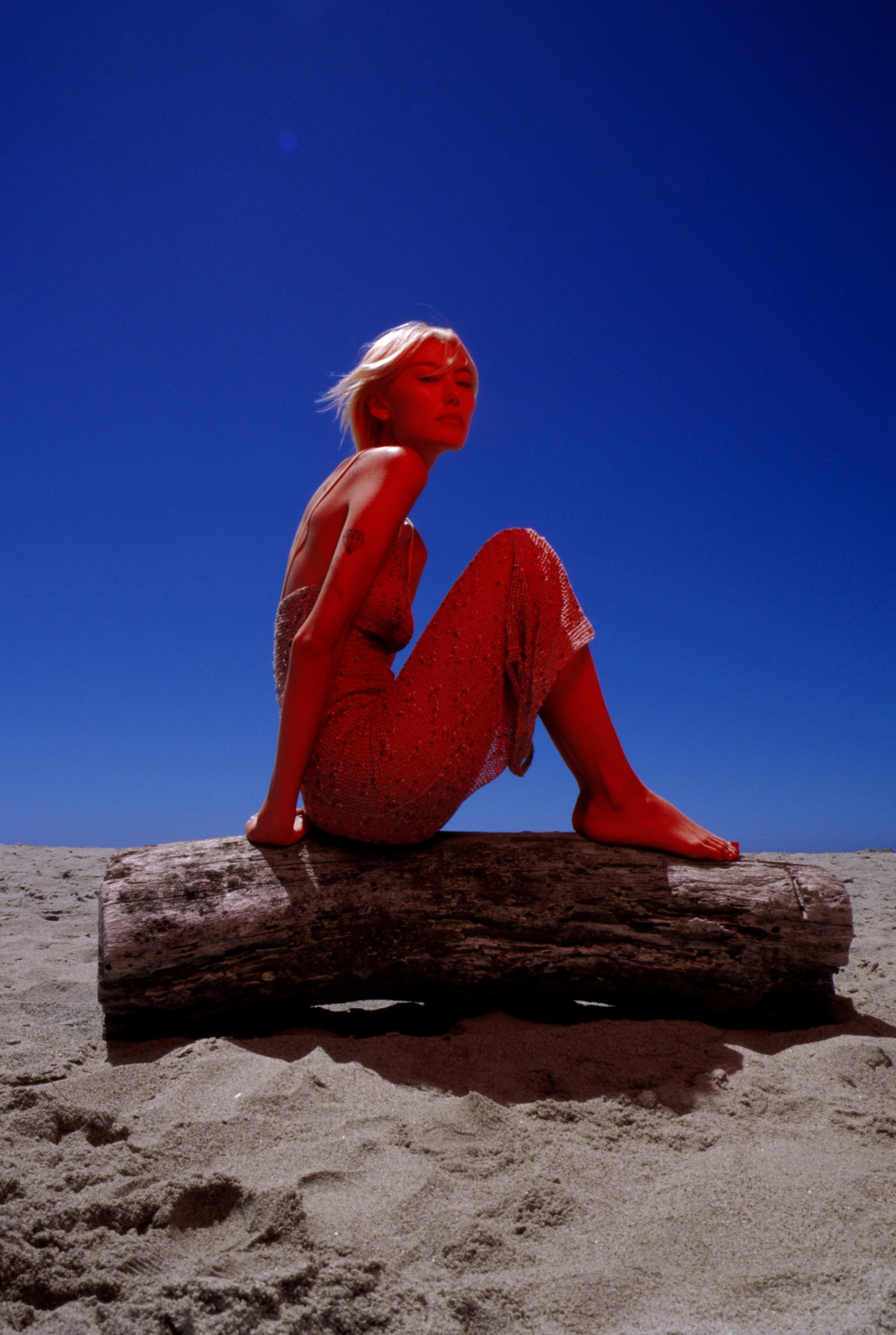 a model cast in red light poses in front of a blue sky, balancing on a wooden log on a sandy floor