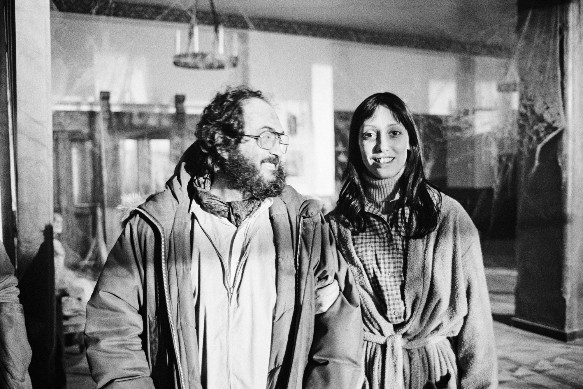 stanley kubrick and shelley duvall stand in the Shining hotel. kubrick looks at duvall smiling, who is smiling down the camera.