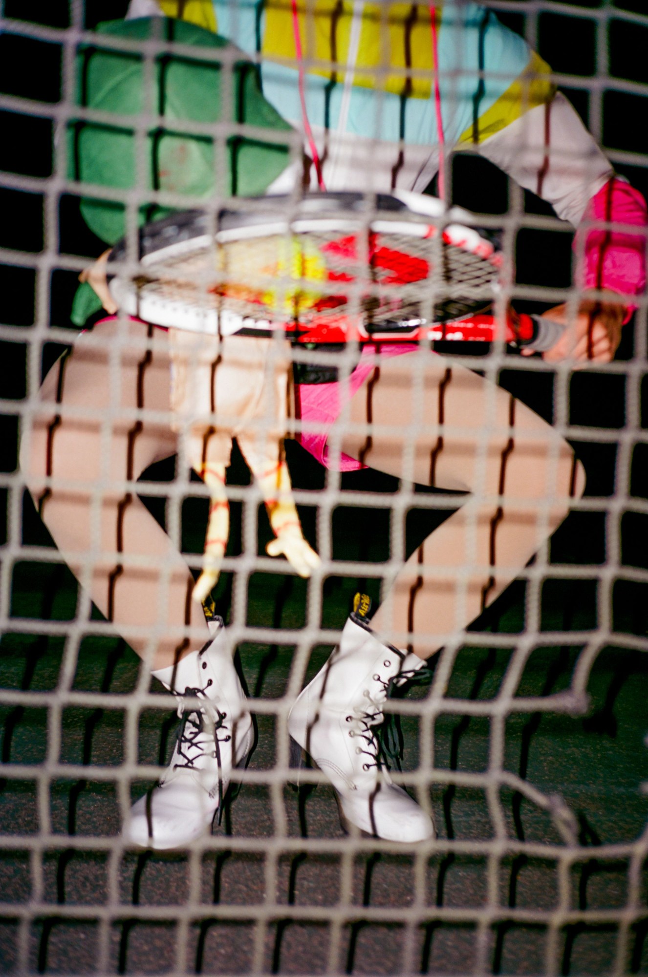 a drag queen in white doc martens and colourful clothes seen from behind tennis net