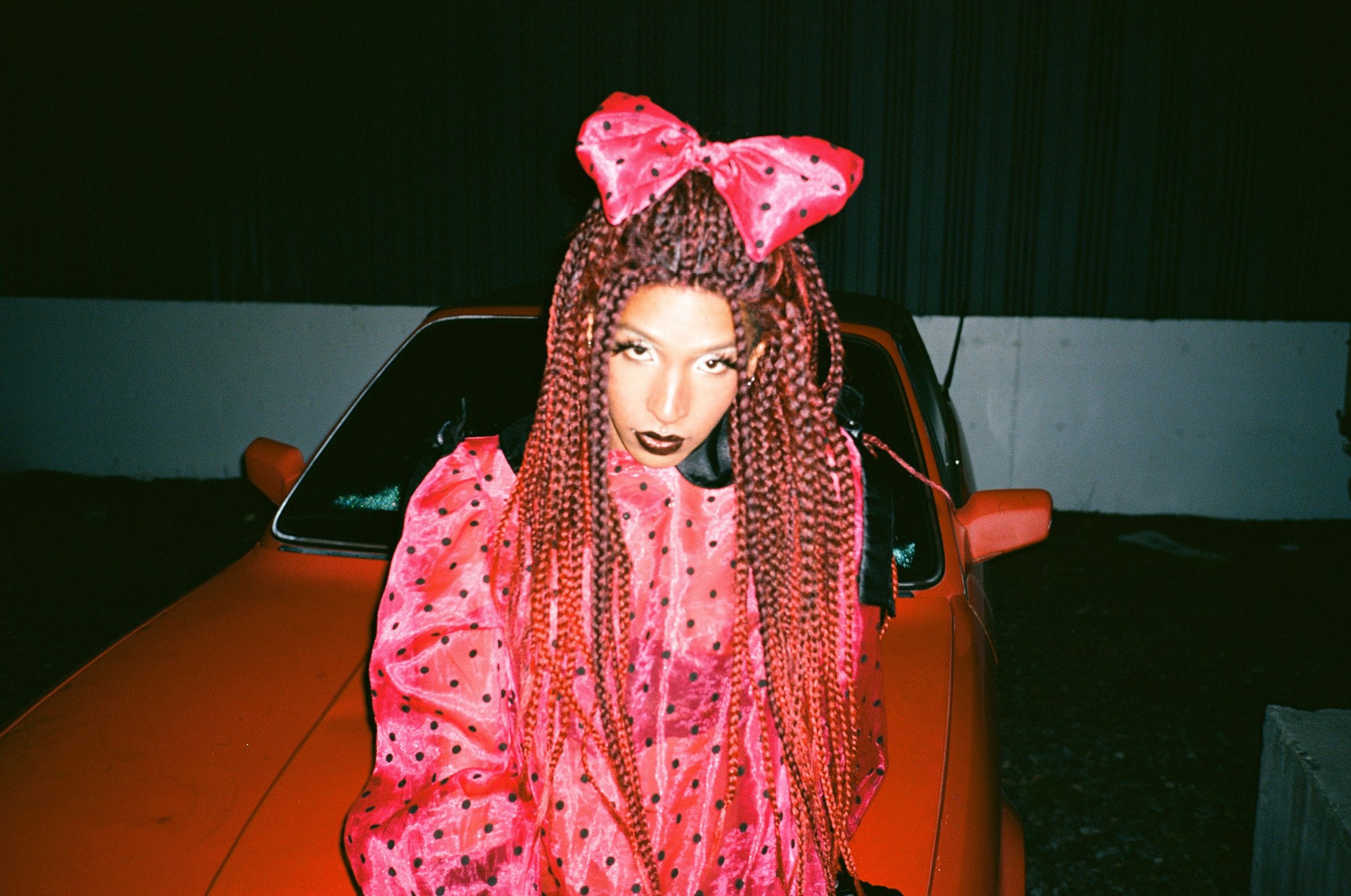 a drag queen in a pink polka dot dress sitting on a red car