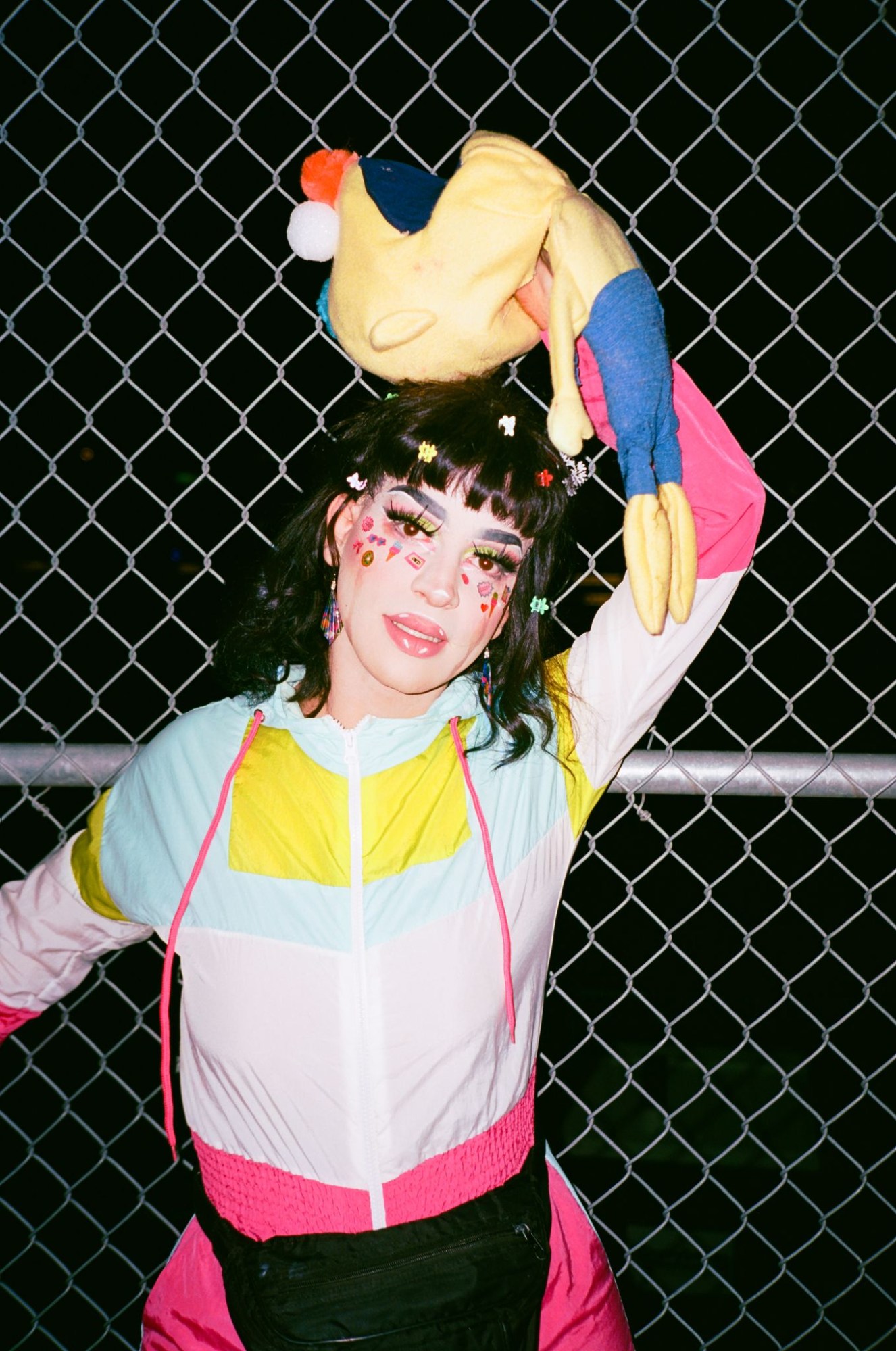 a drag queen in colourful 80s clothing poses in front of a chain fence