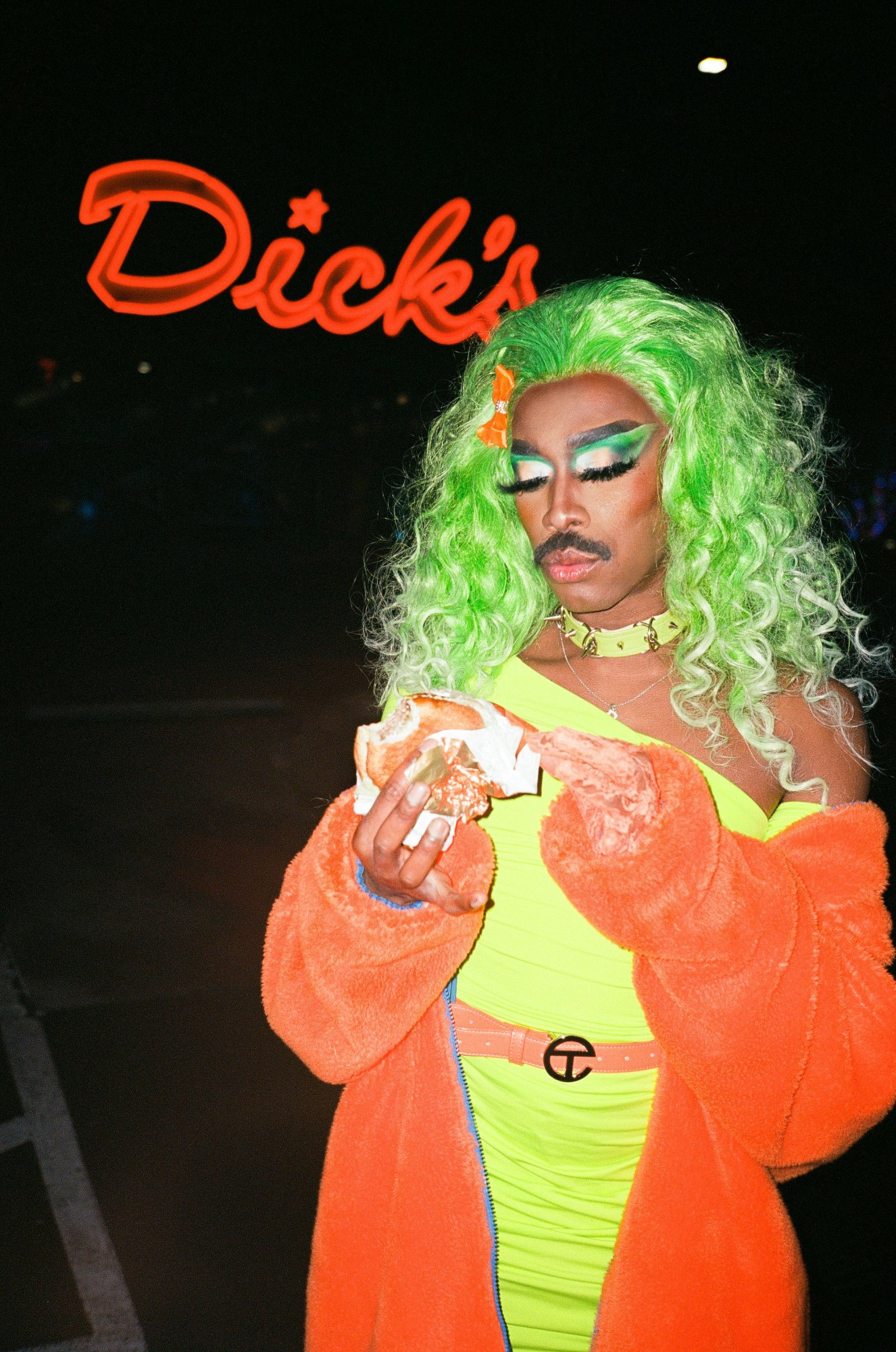 a drag queen eats a burger in front of a sign that says dick's