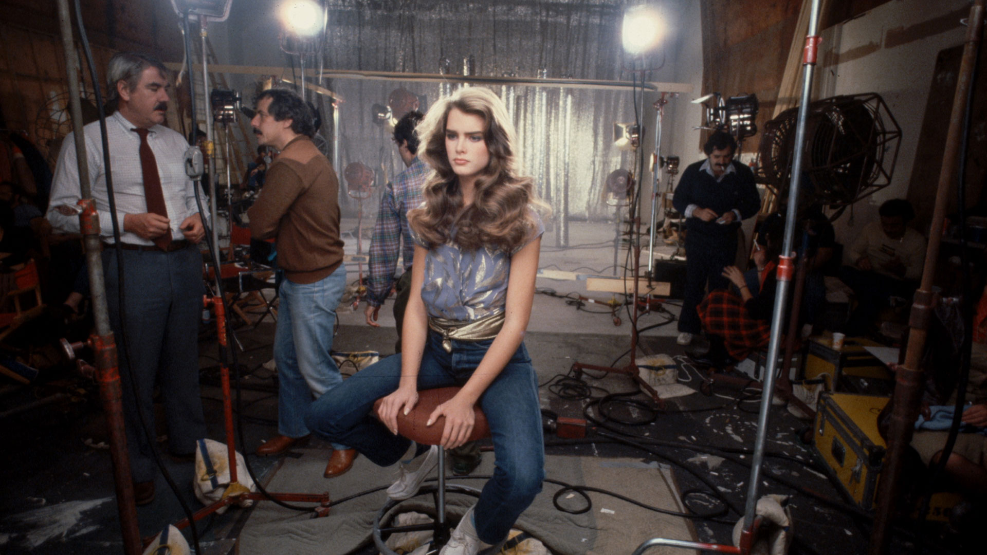 brooke shields sits on a stool in the middle of a busy backstage room