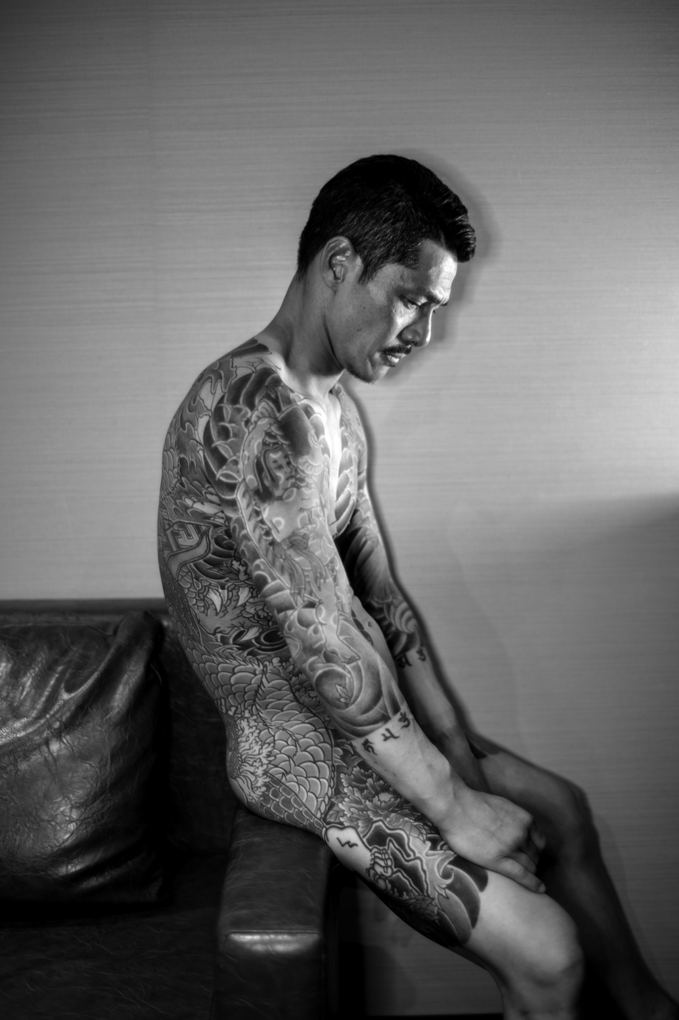 black-and-white image of a naked man shot in profile sitting on a sofa covered in tattoos