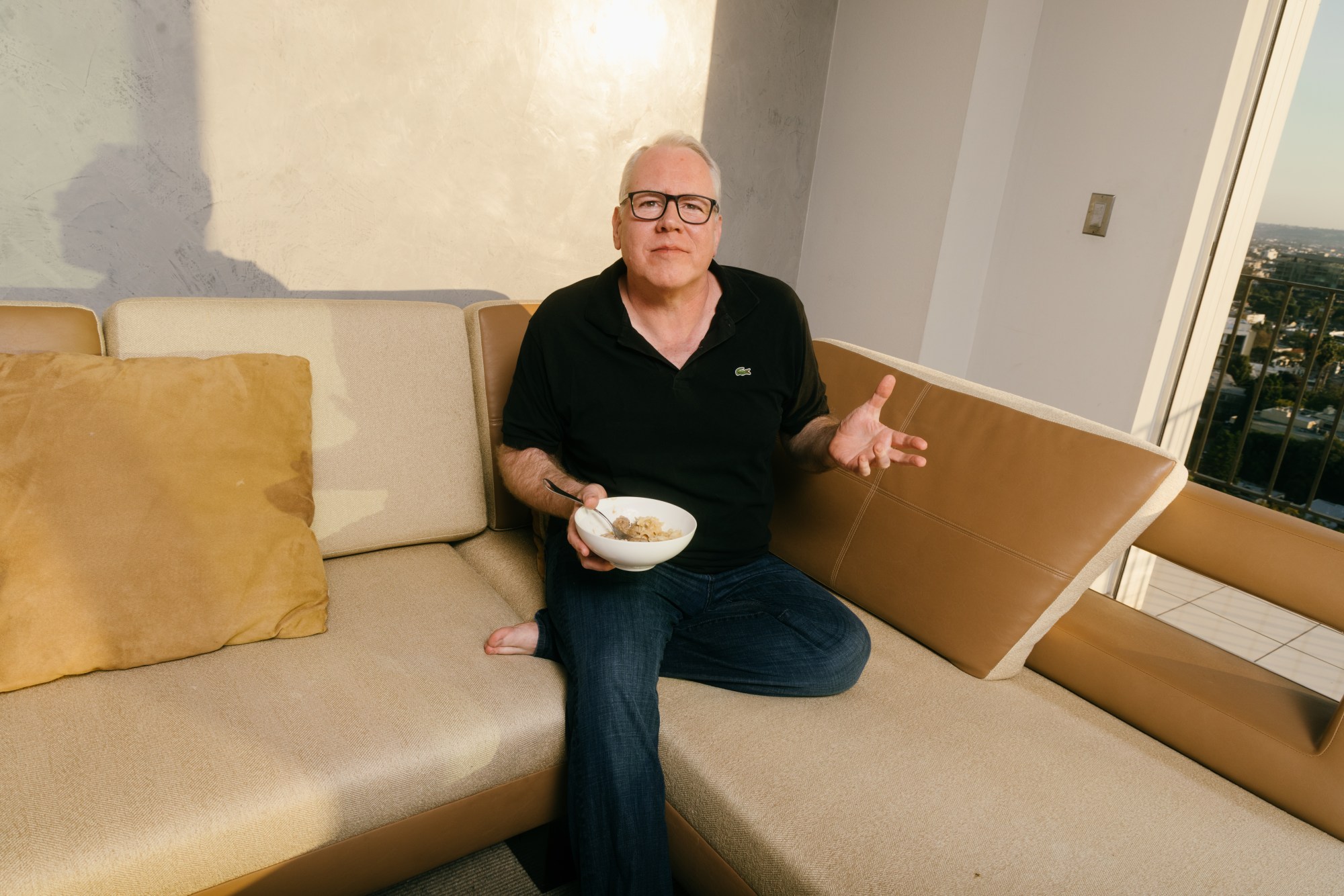 bret easton ellis sits on his couch, gesturing and holding a bowl of cereal