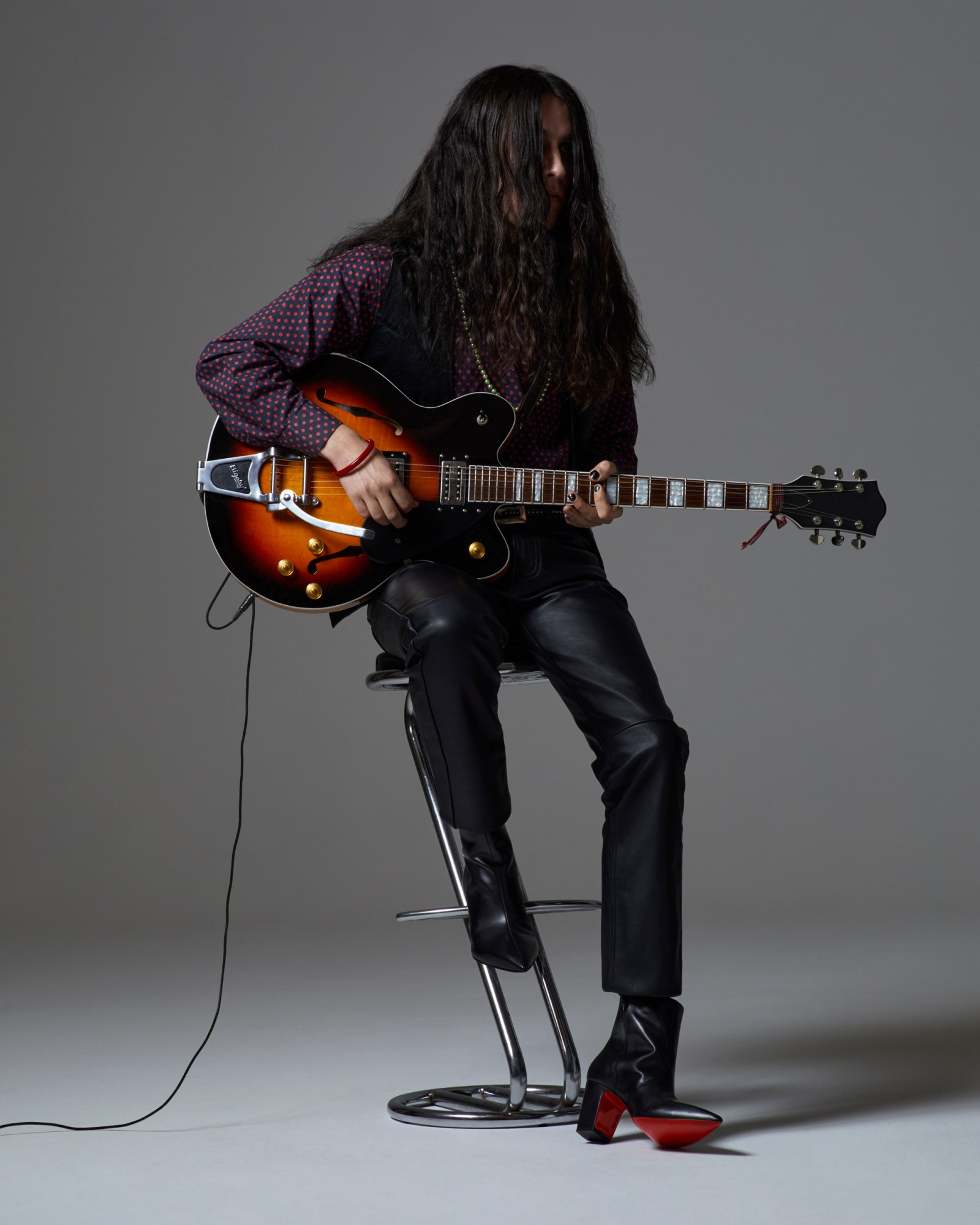 Lewis of Walt Disco wearing a shirt, leather waistcoat and leather trousers and wearing black nappa leather louboutin heel boots while holding a guitar and sitting on a stool