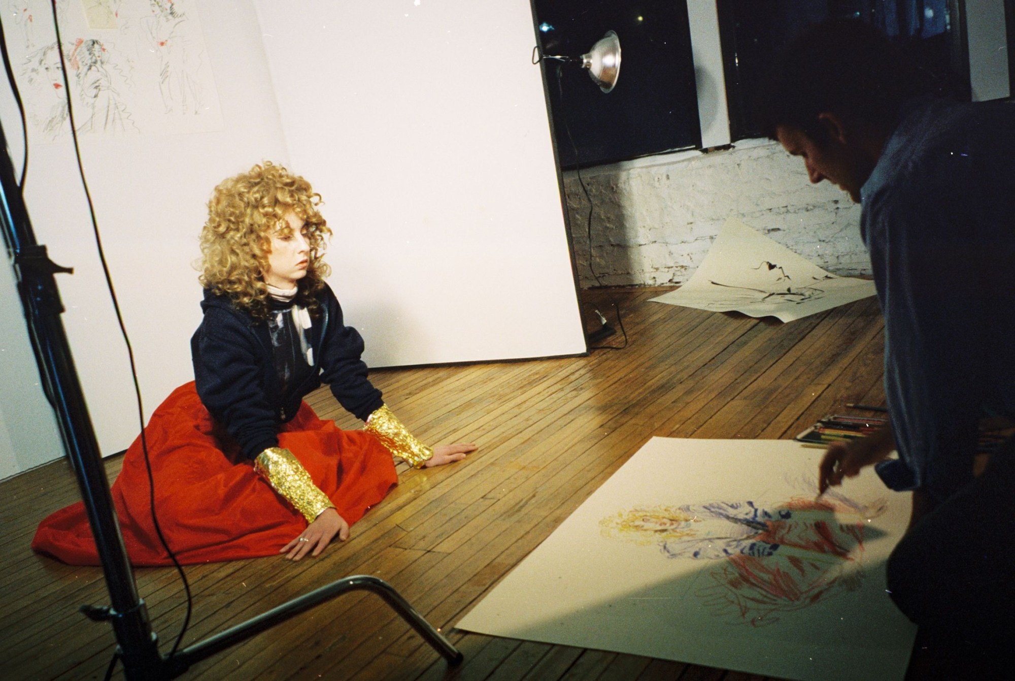 a woman in a black hoodie and red skirt poses for the artist drake carr on the floor of his studio