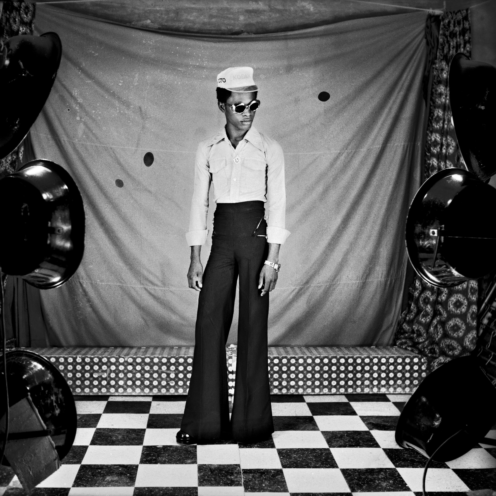 Samuel Fosso wearing black bell bottoms and a cap in his studio.