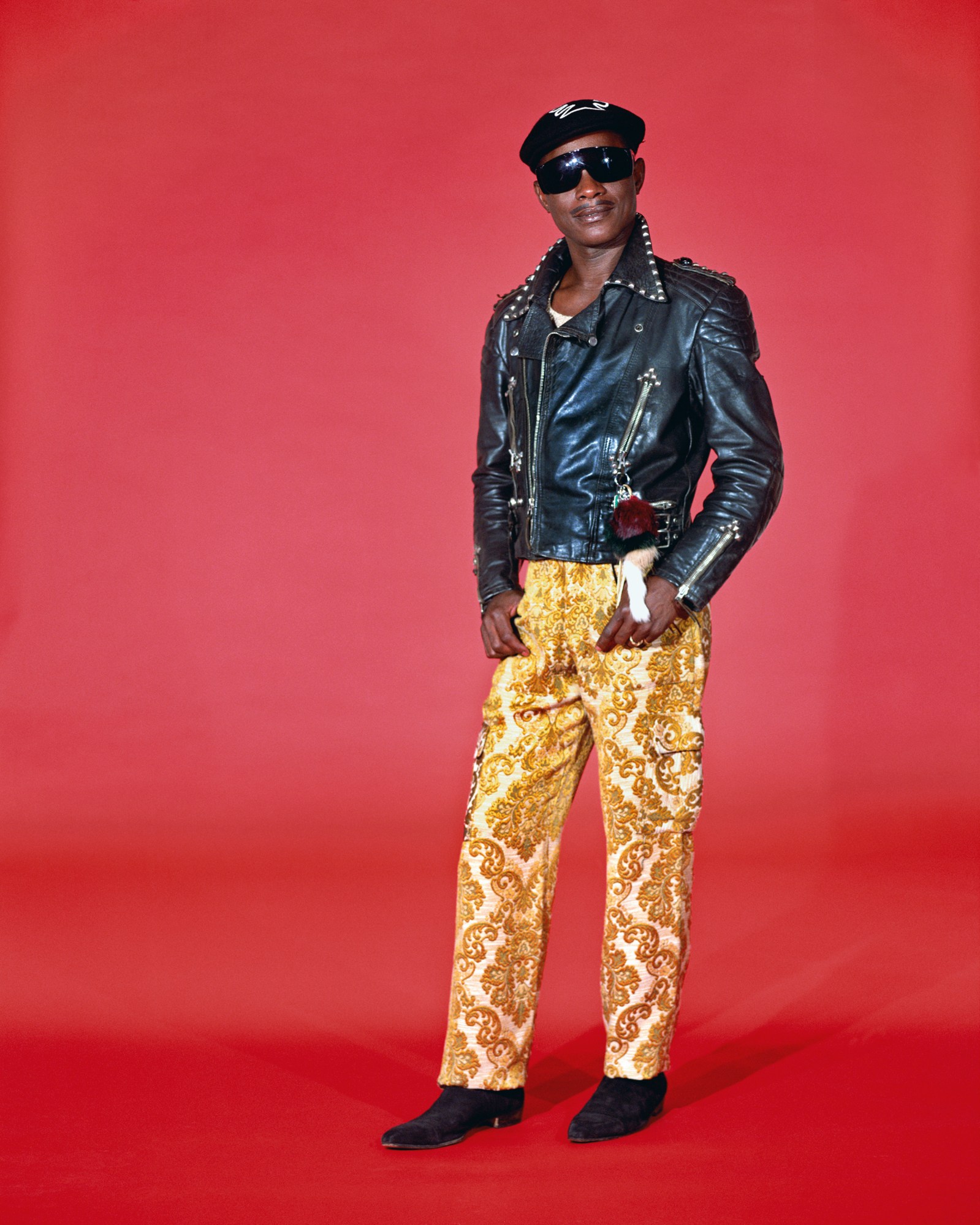 Samuel Fosso wearing black sunglasses, black leather jacket, and jaquard pants in his studio.