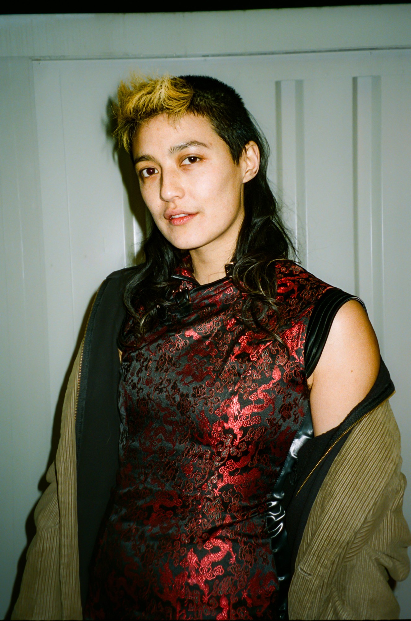 a person wearing a dress with shiny red detailing looks to the camera, their jacket off one shoulder