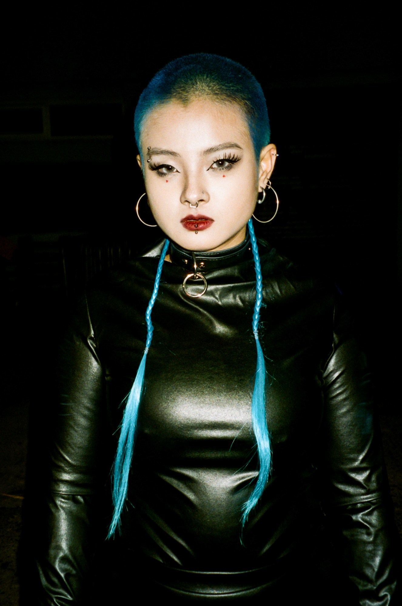 a person with a bright blue shaved head wears a leather one-piece, dog collar and lots of earrings