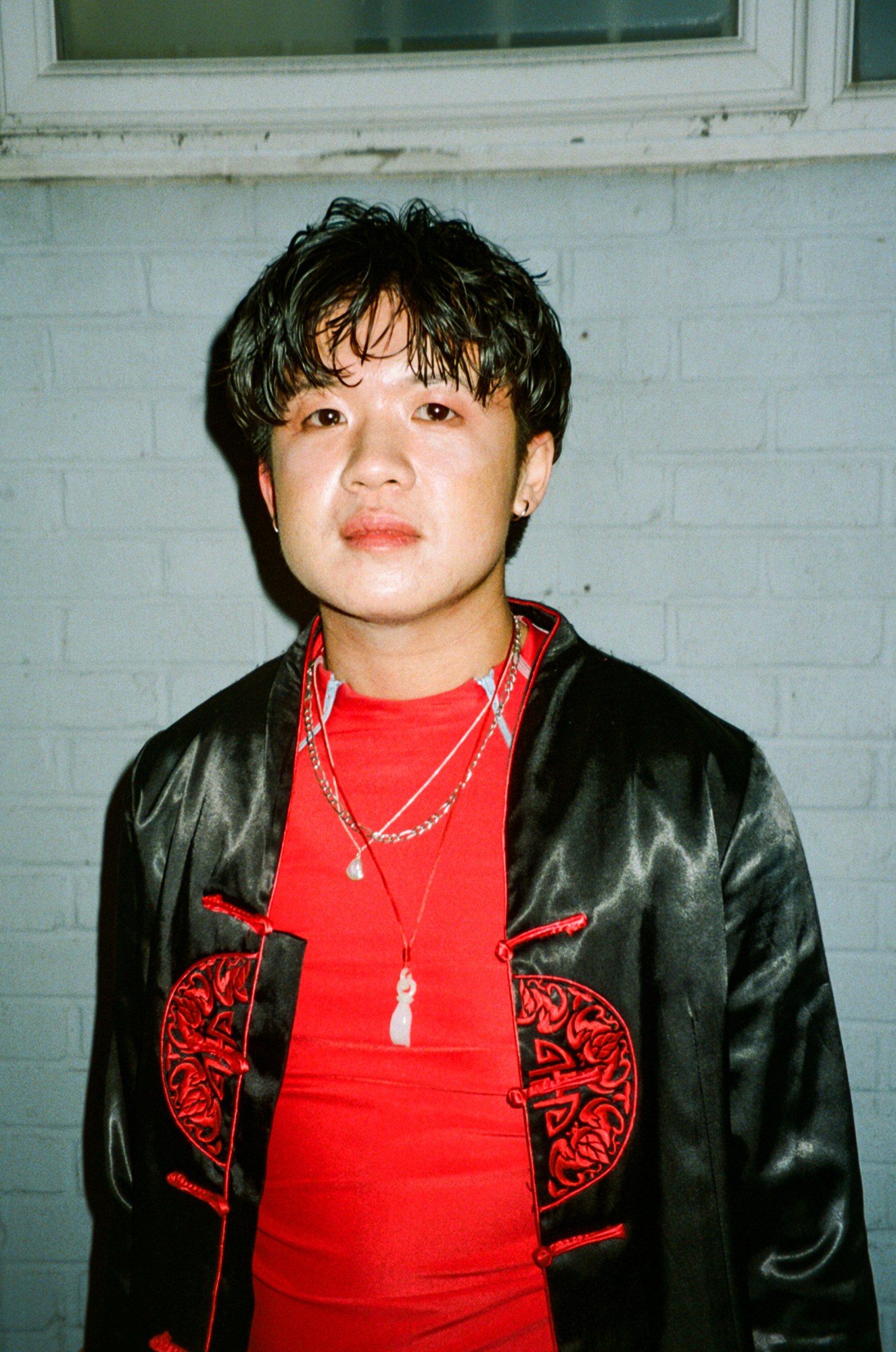 a person in a red top with several necklaces stands against a brick wall and looks to camera