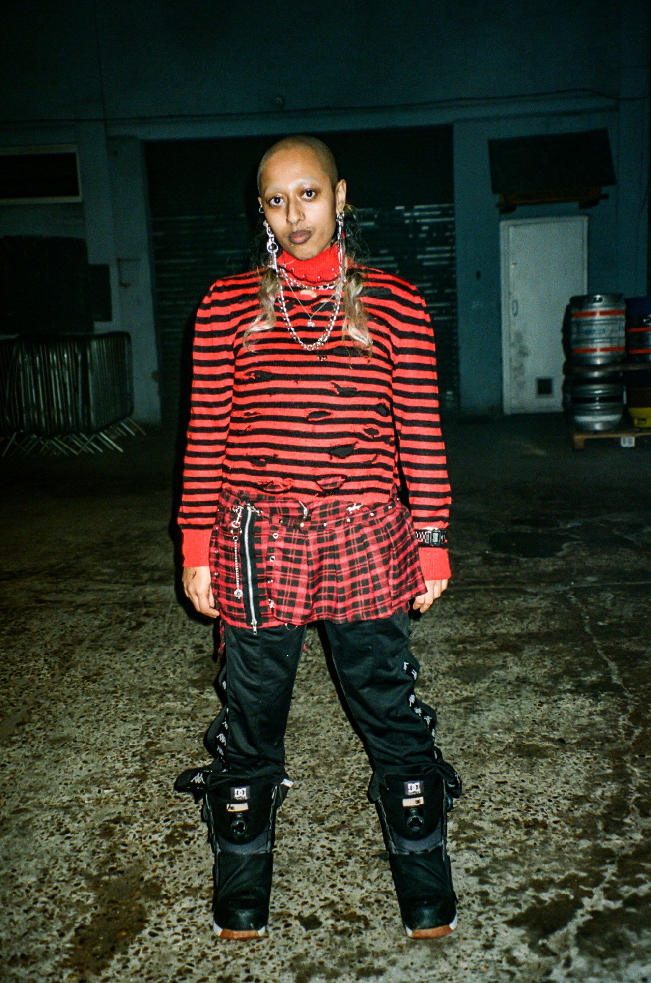 a person with a shaved head and long blonde mullet section poses wearing red and black clothes in a dark yard