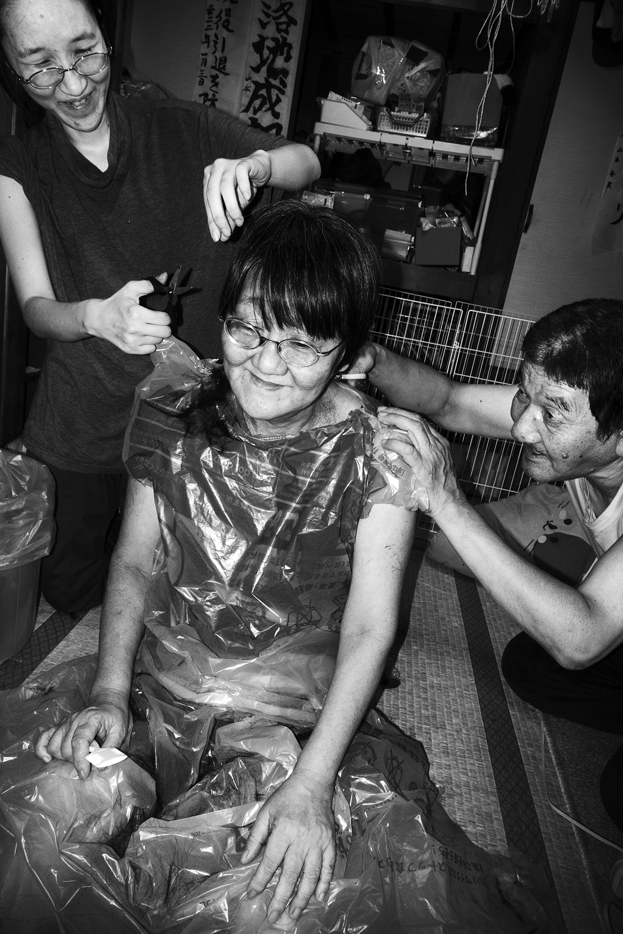 an older japanese woman wears a plastic bag while two family members cut her hair; they're all smiling