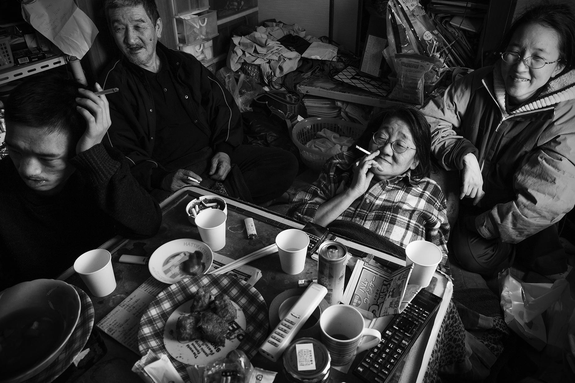 a family sit around a kotatsu table piled with messy plates; they smoke while watching TV