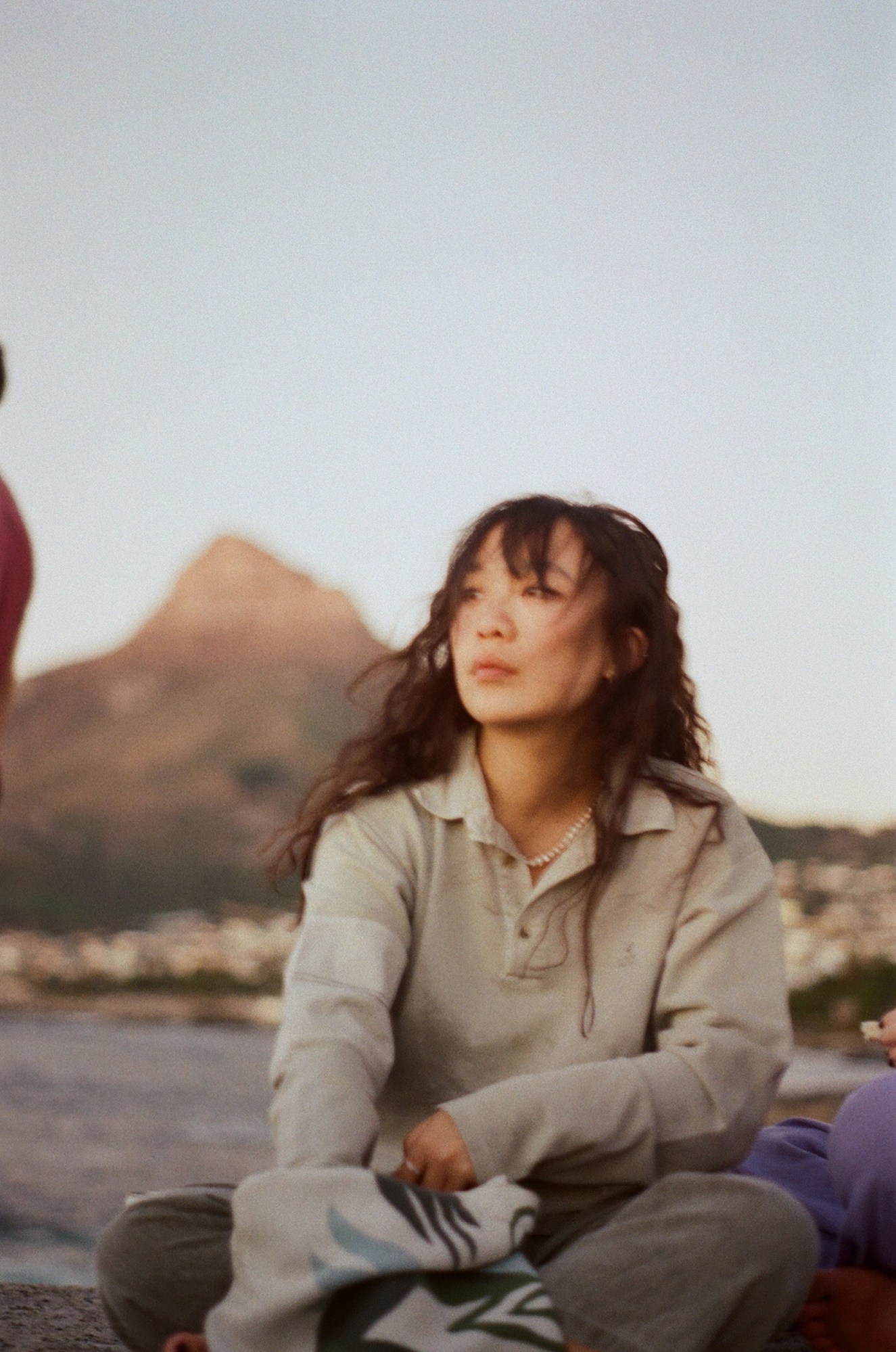 a person with long wavy hair sits crosslegged on a wintry beach, reaching into a tote bag