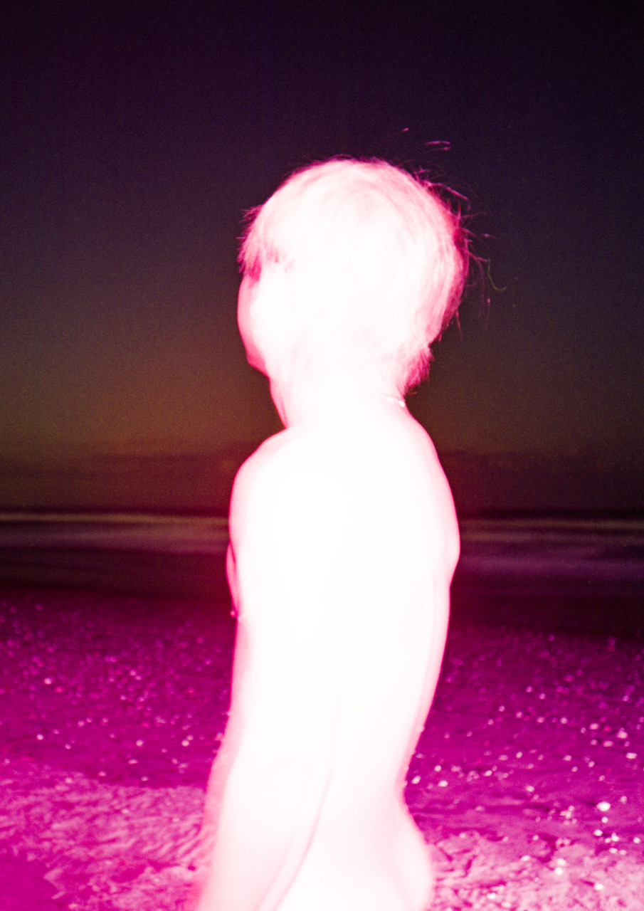 the contrasted silhouette of a nude male body standing on a beach under a purple-pink filter