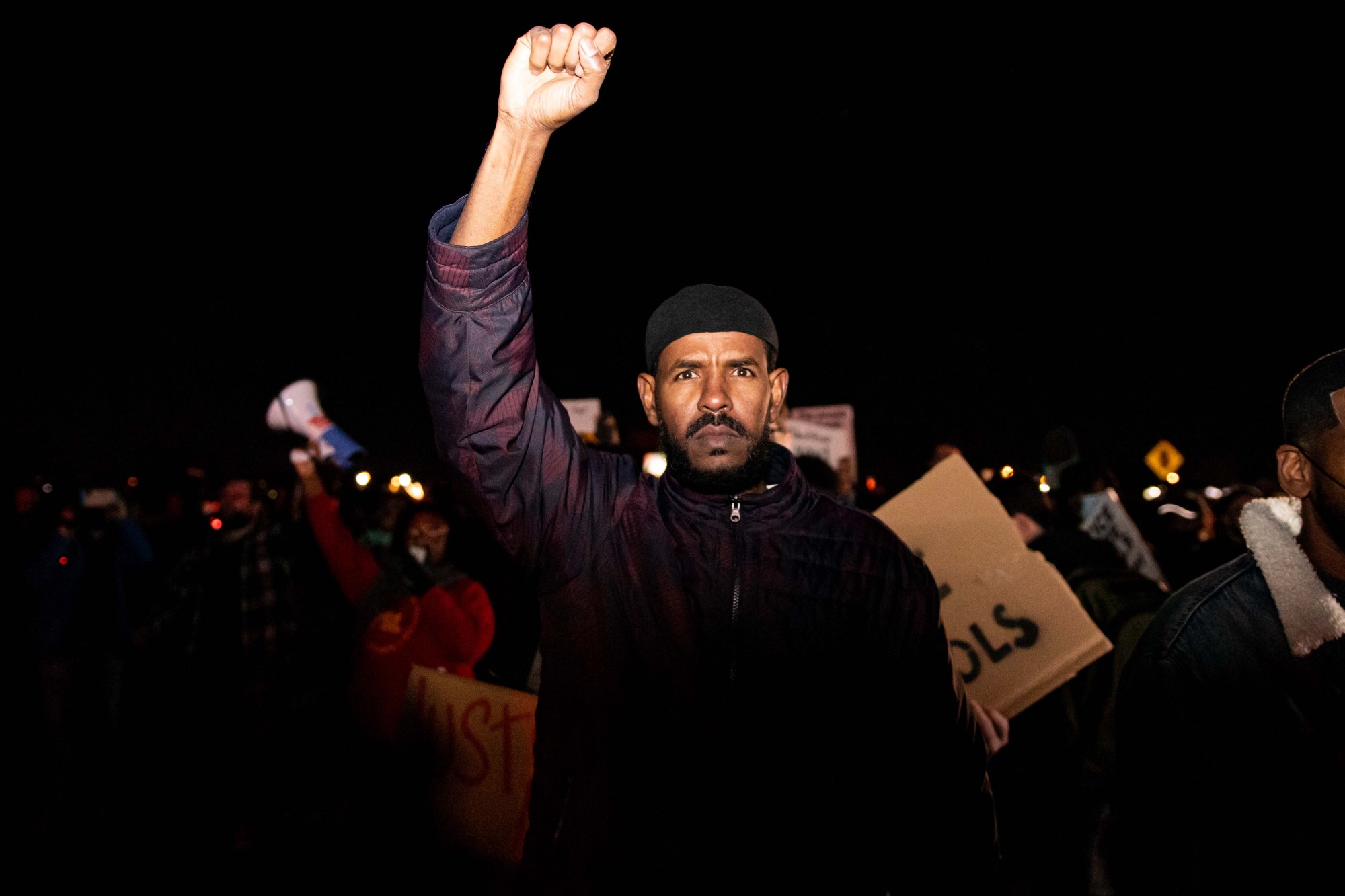 a man with facial hair raises his fist in solidarity; he stands in a crowd of nighttime protesters