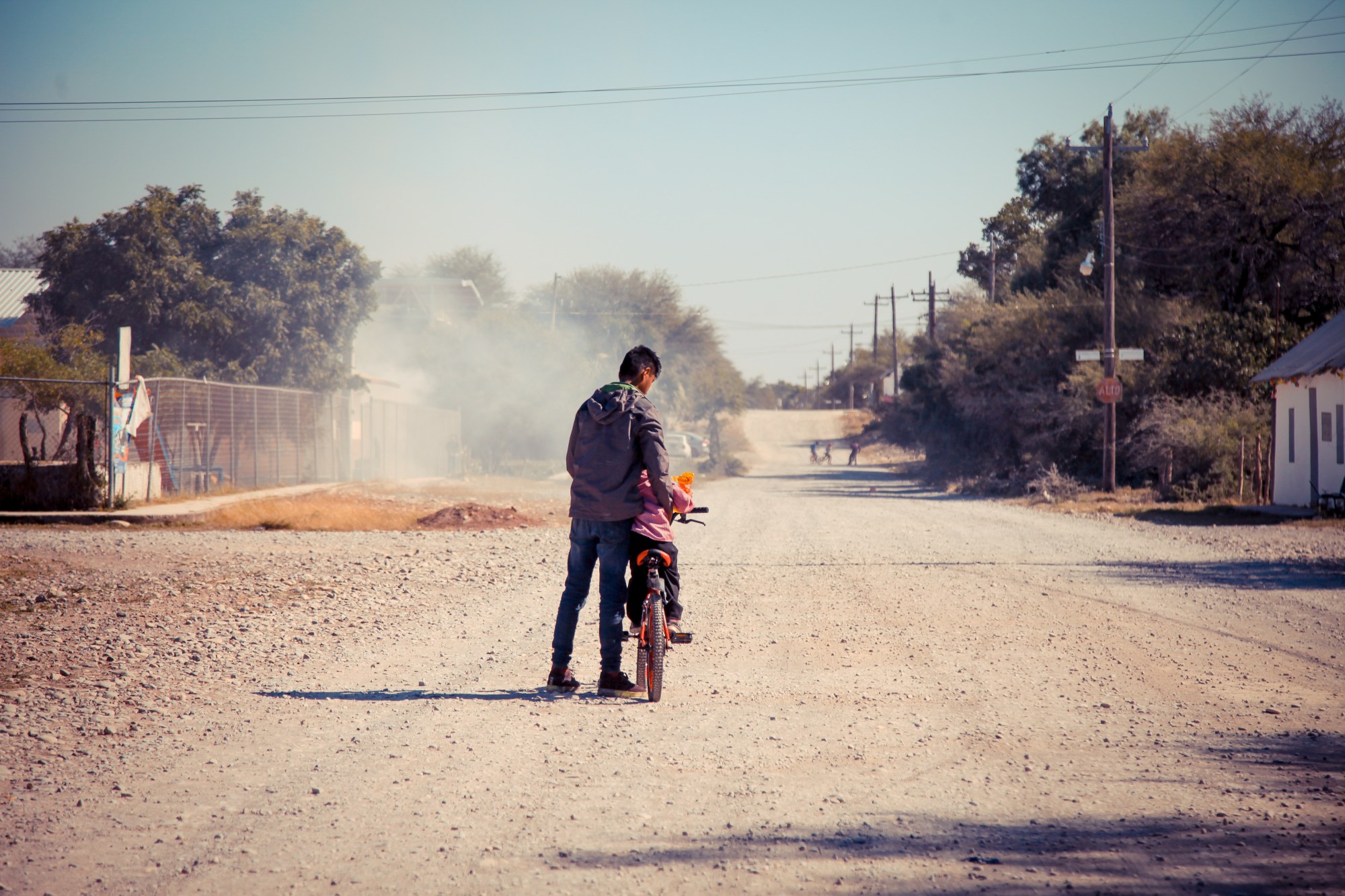 a older child holds their young sibling upright on a bicycle on a dusty road