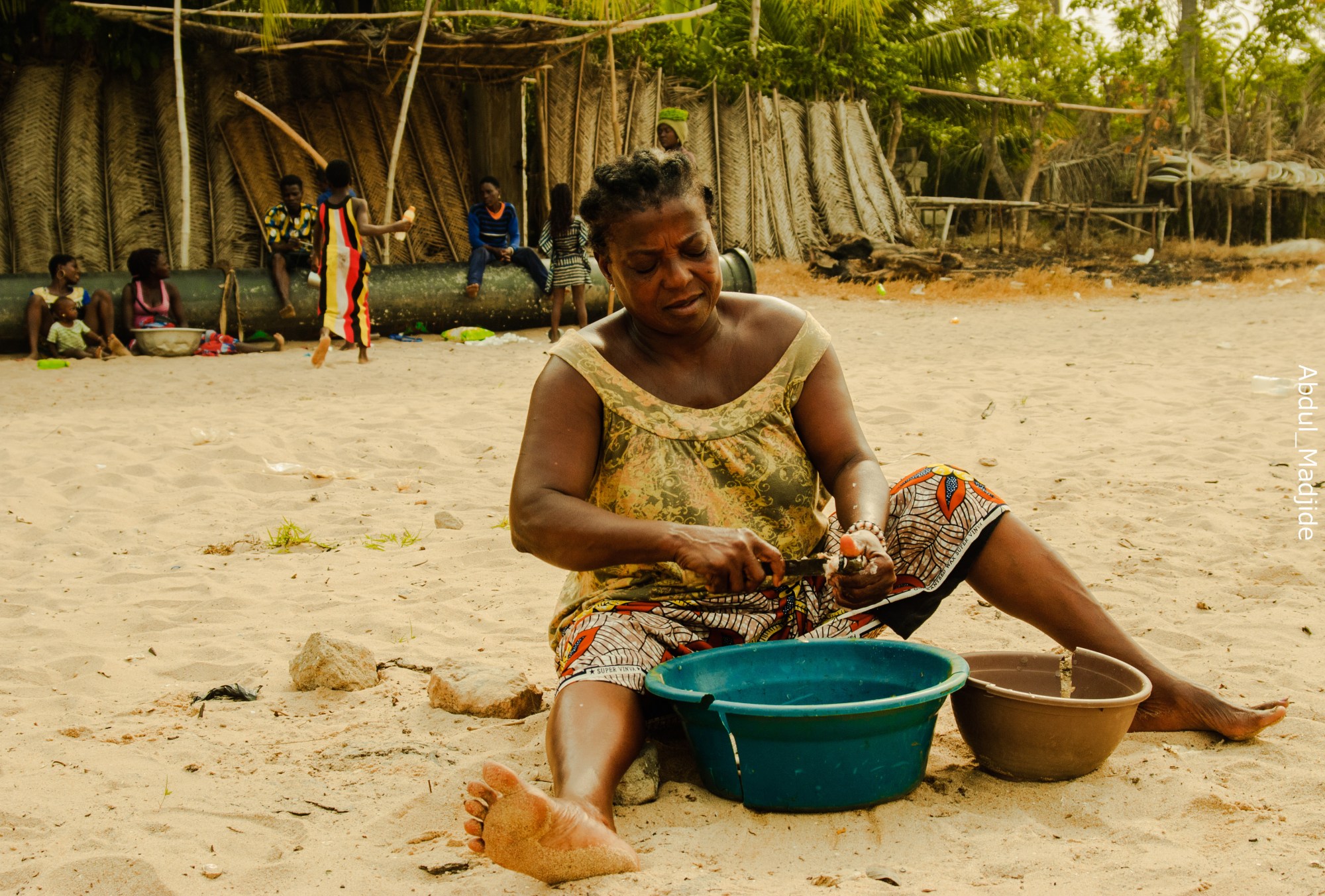 a woman in a patterned dress sits on a beach removing fish scales over two plastic tubs