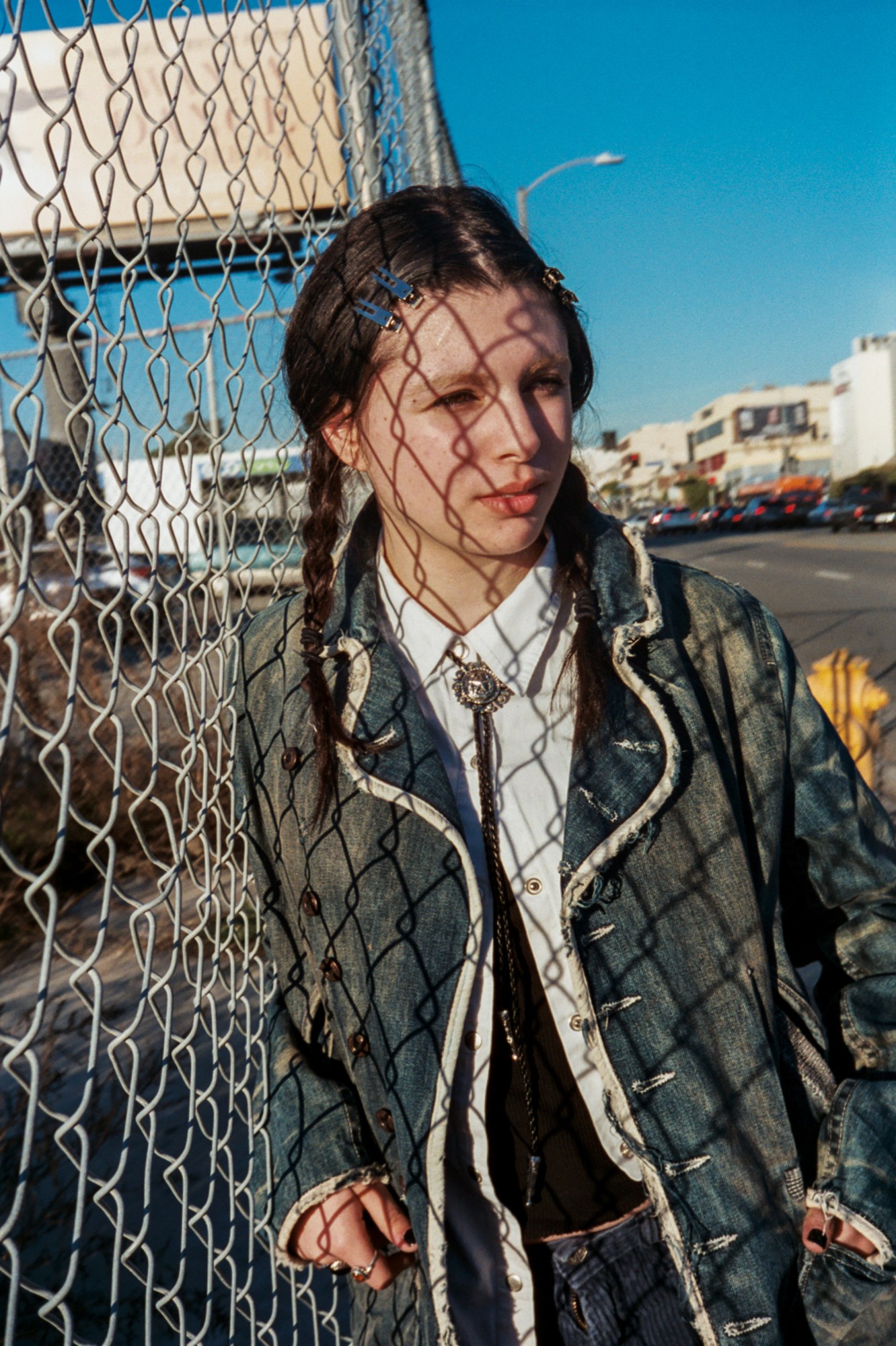 a girl wearing a long denim coat and pigtails stands against a chain link fence