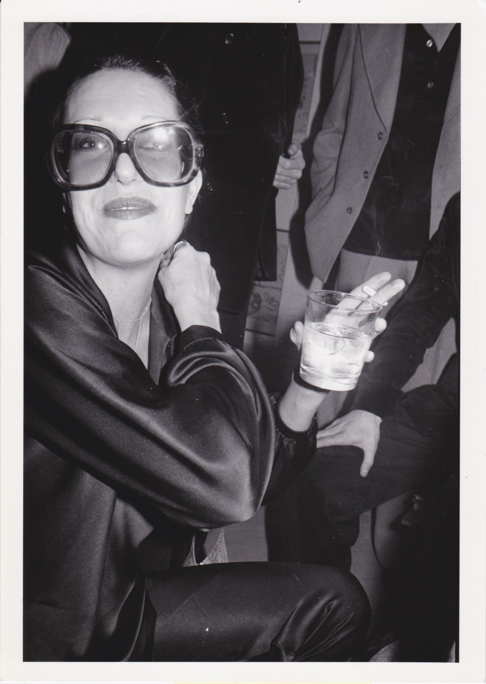 a woman in oversized sunglasses and a silk suit holds a drink and cigarette in one hand