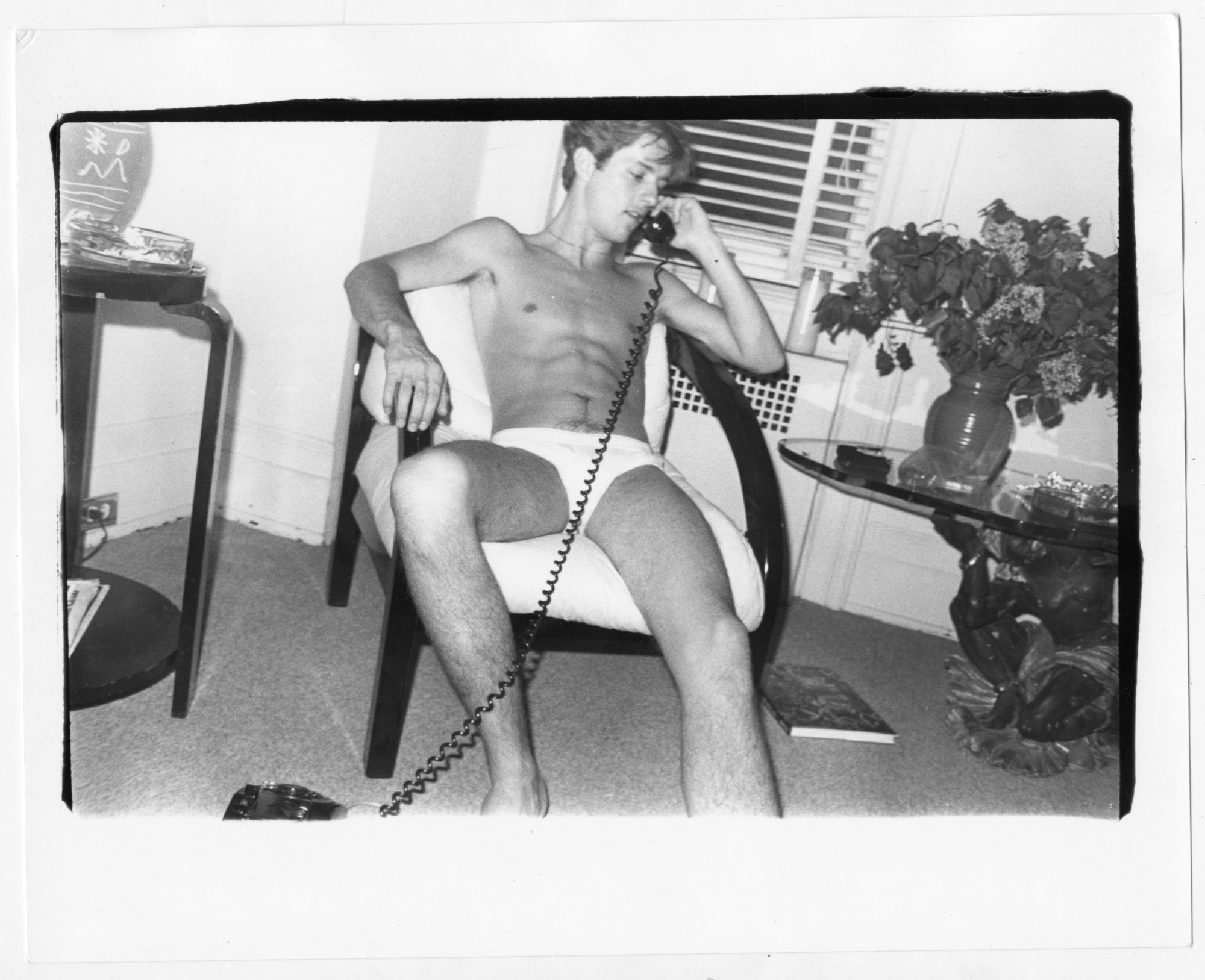 a young man in white briefs sits in a hotel room hair talking on a wired phone