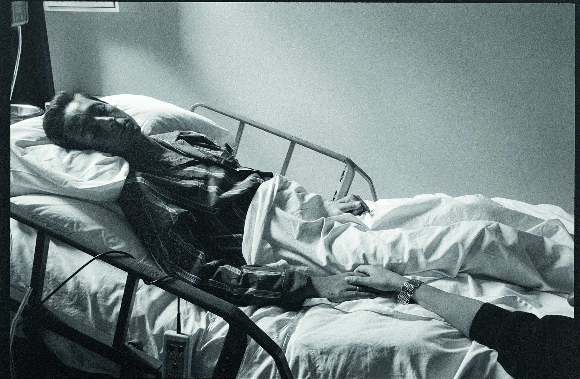 Exclusive photograph from Gideon Mendel's exhibition 'The Ward Revisited' at The Fitzrovia Chapel of a man lying in a hospital bed holding someone's hand while in Broderip AIDS ward in Middlesex Hospital.