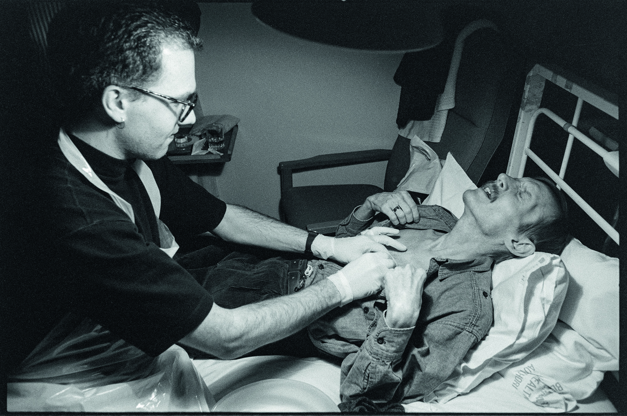 Photograph from Gideon Mendel's exhibition 'The Ward Revisited' at The Fitzrovia Chapel of a man in a hospital bed in Broderip AIDS ward in Middlesex Hospital being examined by a doctor