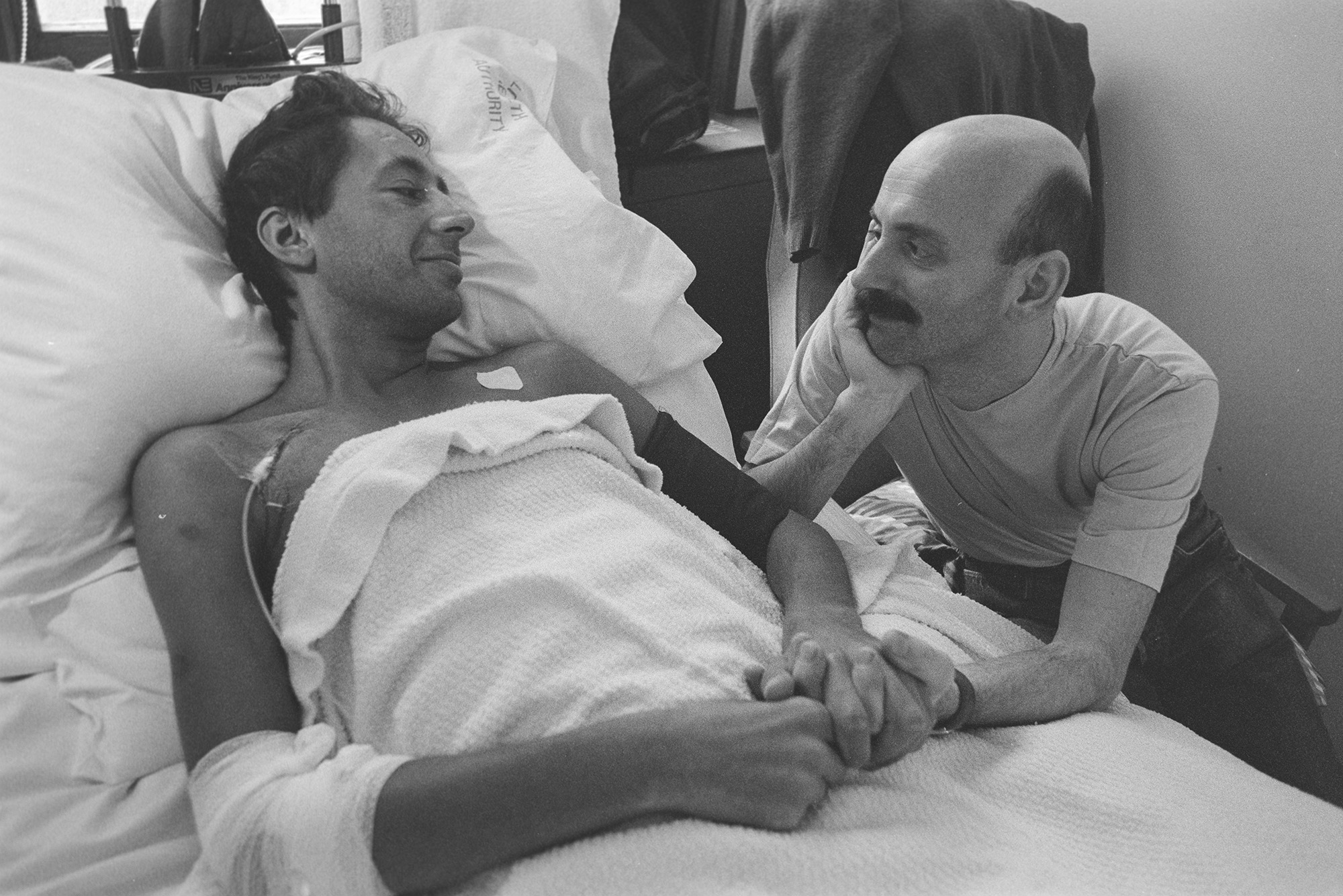 Photograph from Gideon Mendel's exhibition 'The Ward Revisited' at The Fitzrovia Chapel of a man lying in a hospital bed with their partner by their side in Broderip AIDS ward in Middlesex Hospital.