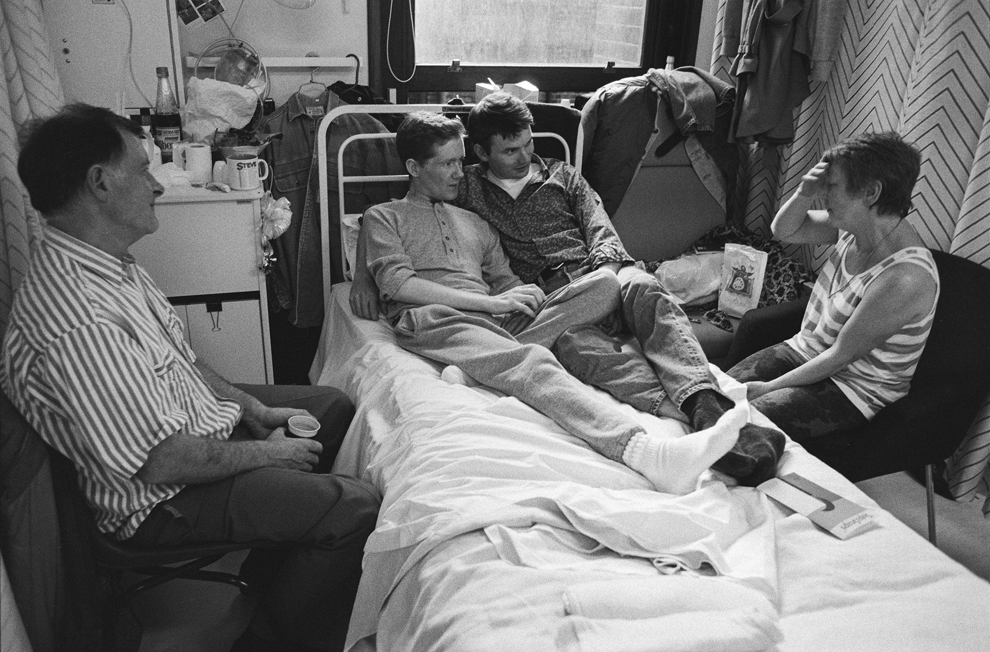 Photograph from Gideon Mendel's exhibition 'The Ward Revisited' at The Fitzrovia Chapel of two men lying on a hospital bed in Broderip AIDS ward in Middlesex Hospital while an older woman and man sit by them.