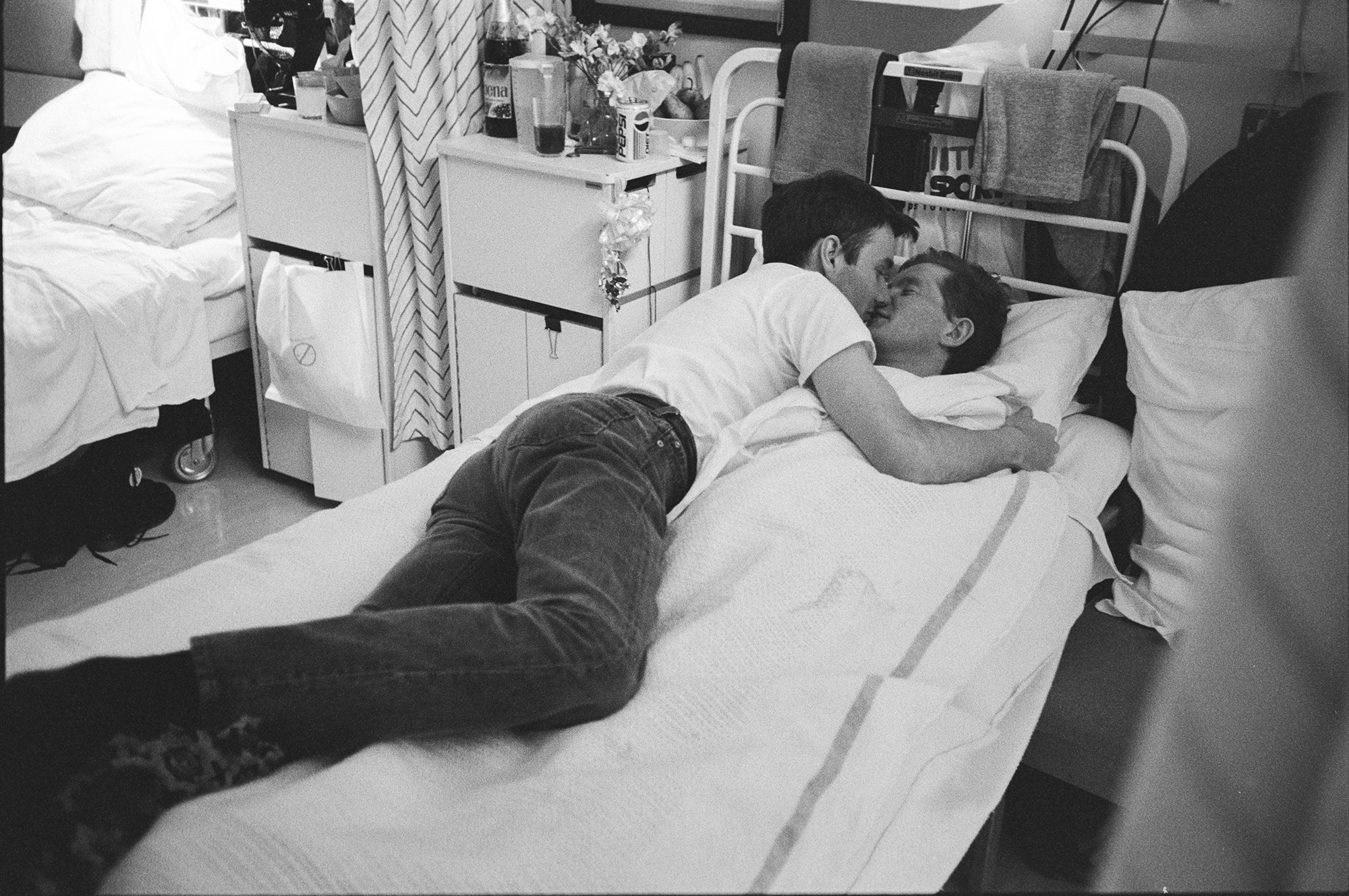 Photograph from Gideon Mendel's exhibition 'The Ward Revisited' at The Fitzrovia Chapel of two men kissing in a hospital bed in Broderip AIDS ward in Middlesex Hospital