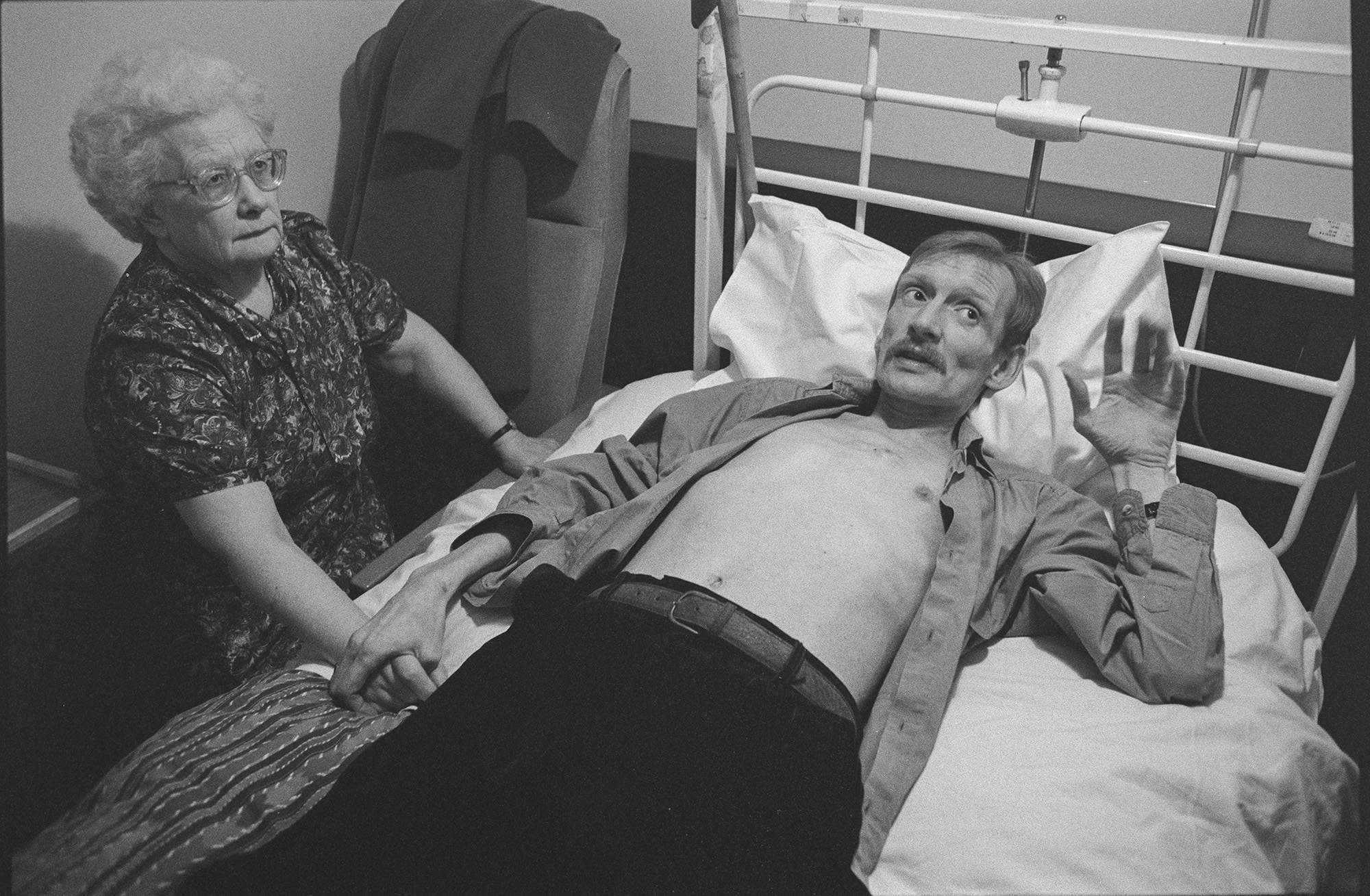 Photograph from Gideon Mendel's exhibition 'The Ward Revisited' at The Fitzrovia Chapel of a man lying in a hospital bed in Broderip AIDS ward in Middlesex Hospital while on old lady holds his hand.