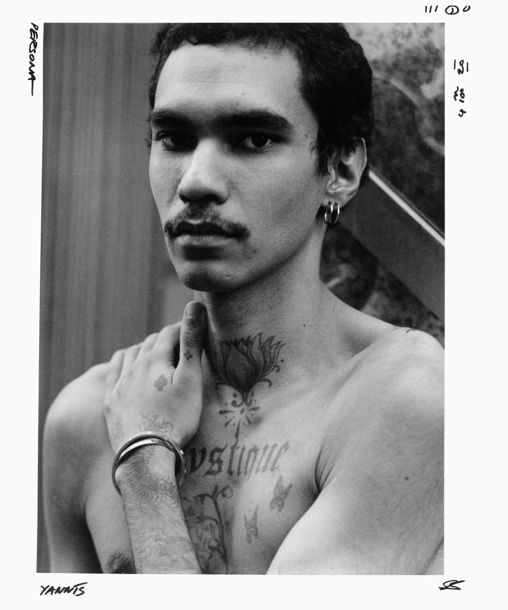 a portrait of a topless man holding his arm up to his shoulder with tattoos, bracelets and earrings