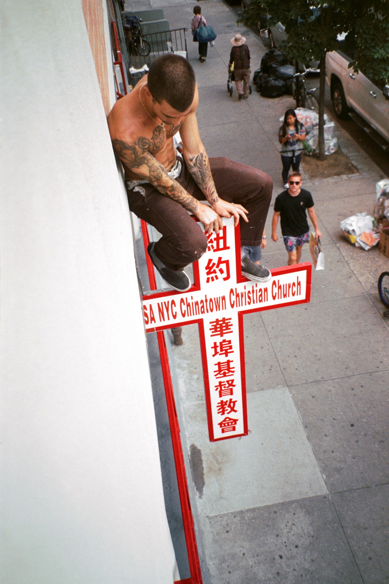 a man with tattoos sitting on top of a cross sign for nyc chinatown christian church