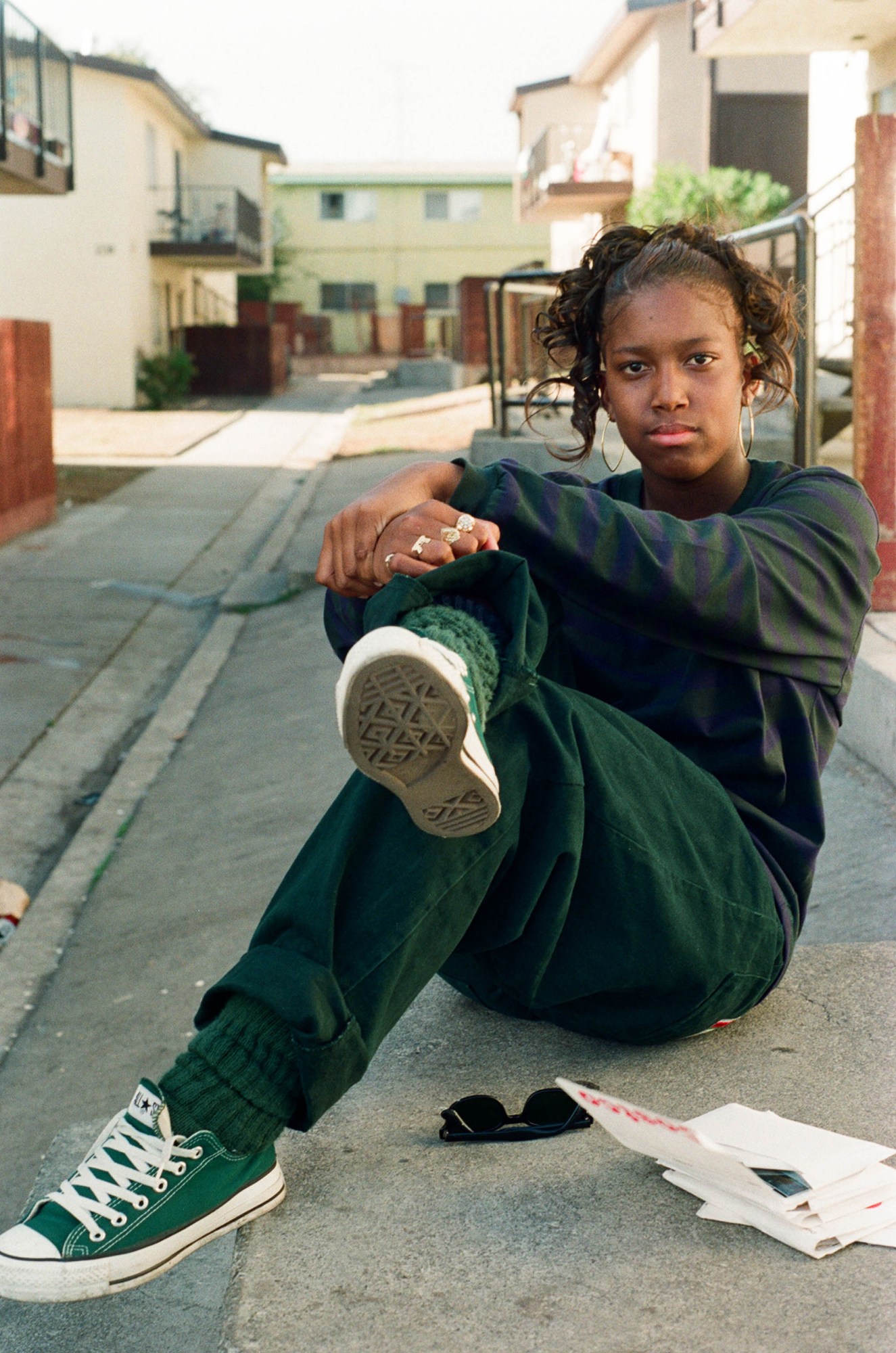 A Black woman with curly hair, hoop earrings, and rings sitting on the curb in green Converse sneakers.